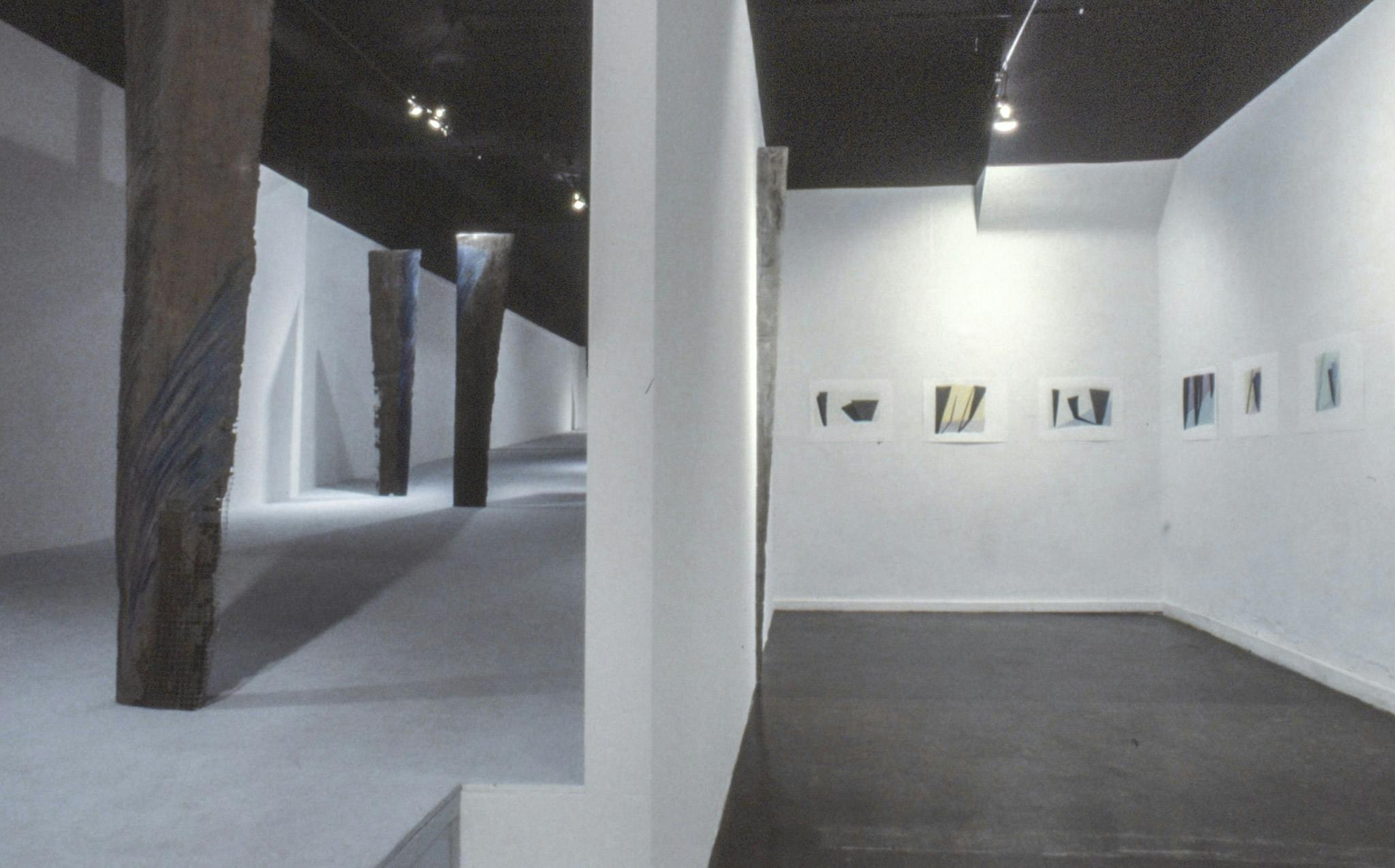 A gallery space divided into two visible sections. One section is an elevated platform with vertical cement fondu planks. The other section has a black floor and abstract paintings on the white walls. 