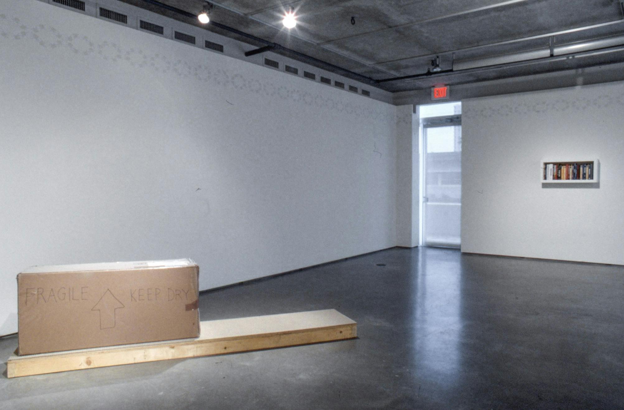 A large sculpture is installed in a gallery. The sculpture is a sealed large cardboard box placed on a piece of flat wood. Words such as Fragile and Keep Dry are written on the side of the box.