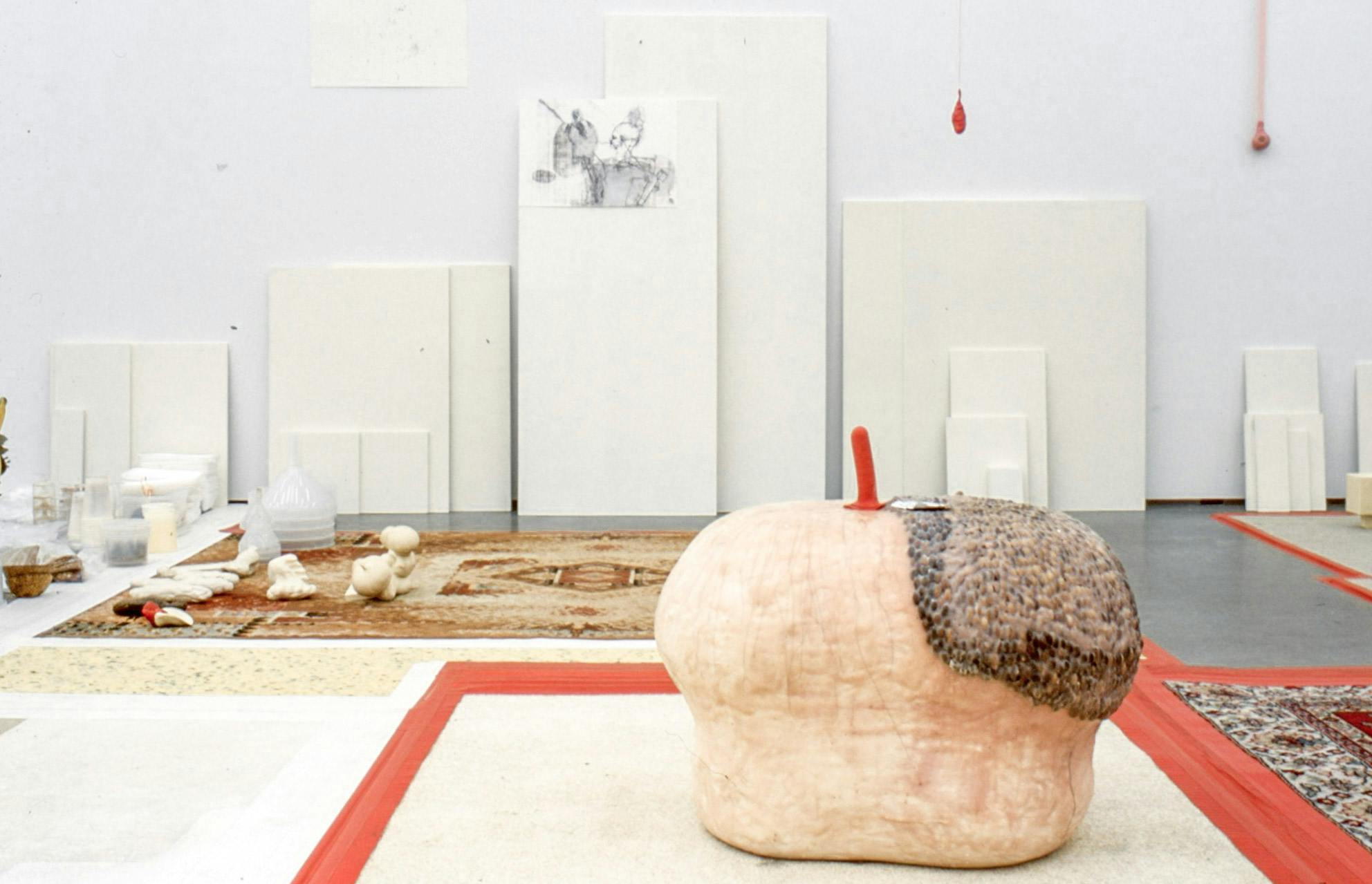 Installation image of artworks in a gallery. A variety of found objects, including shellfish and a toy shovel are placed on a partially carpeted floor. Various sized white planks lean against the far wall.