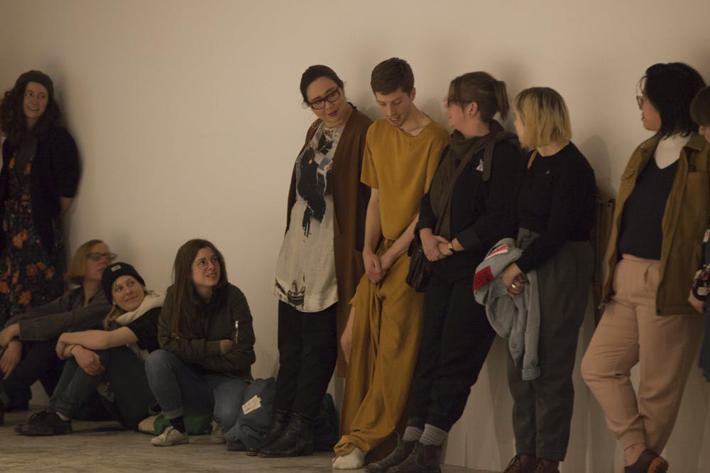 A performer dressed in yellow stands with audience members by the wall of a gallery space. Five audience members are visibly standing on the left side of the performer, and three on the right. 