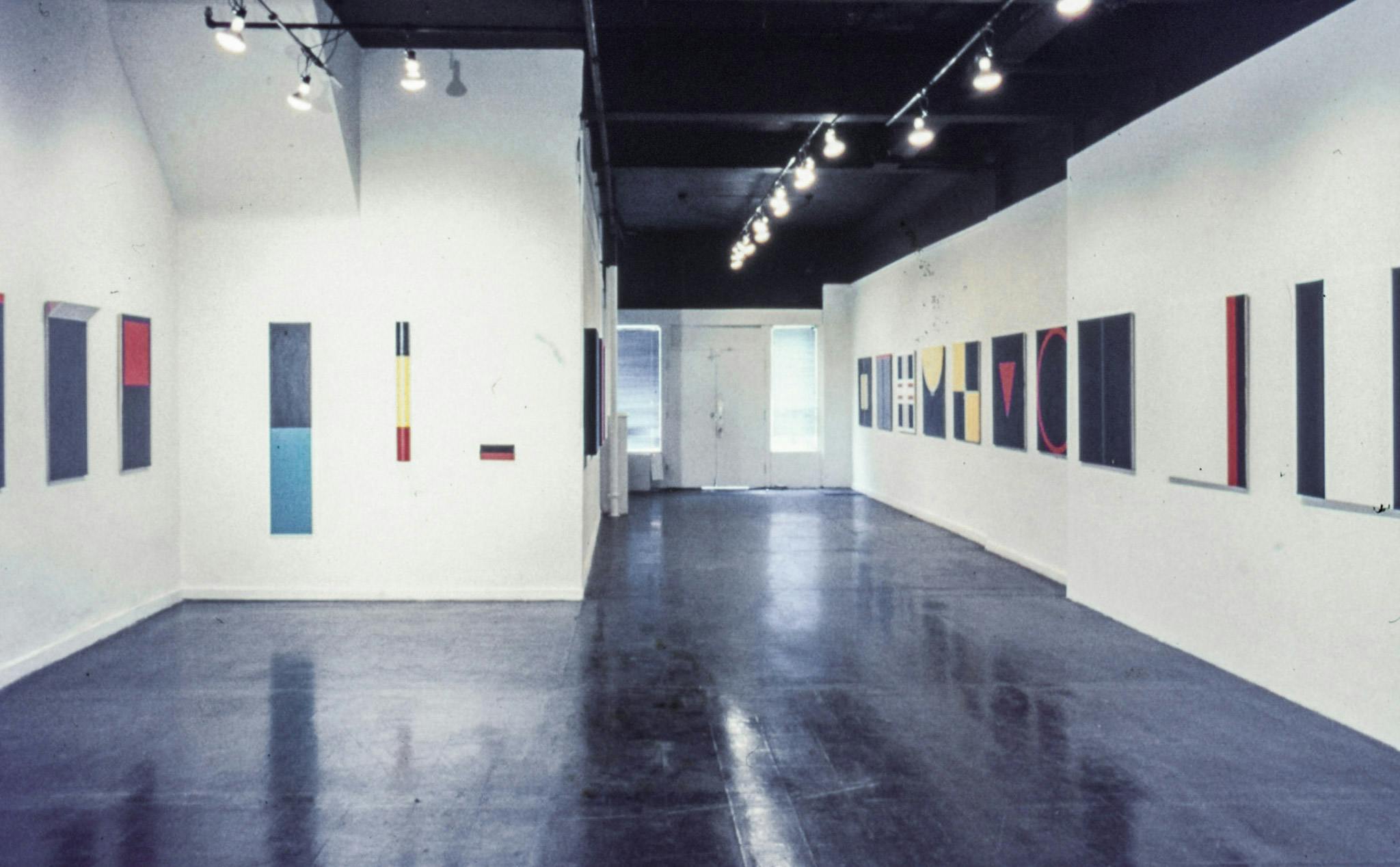 Several abstract paintings line the walls of a gallery. Most of the paintings are on square canvases, and show geometric shapes in blue, yellow, white, black, and red.