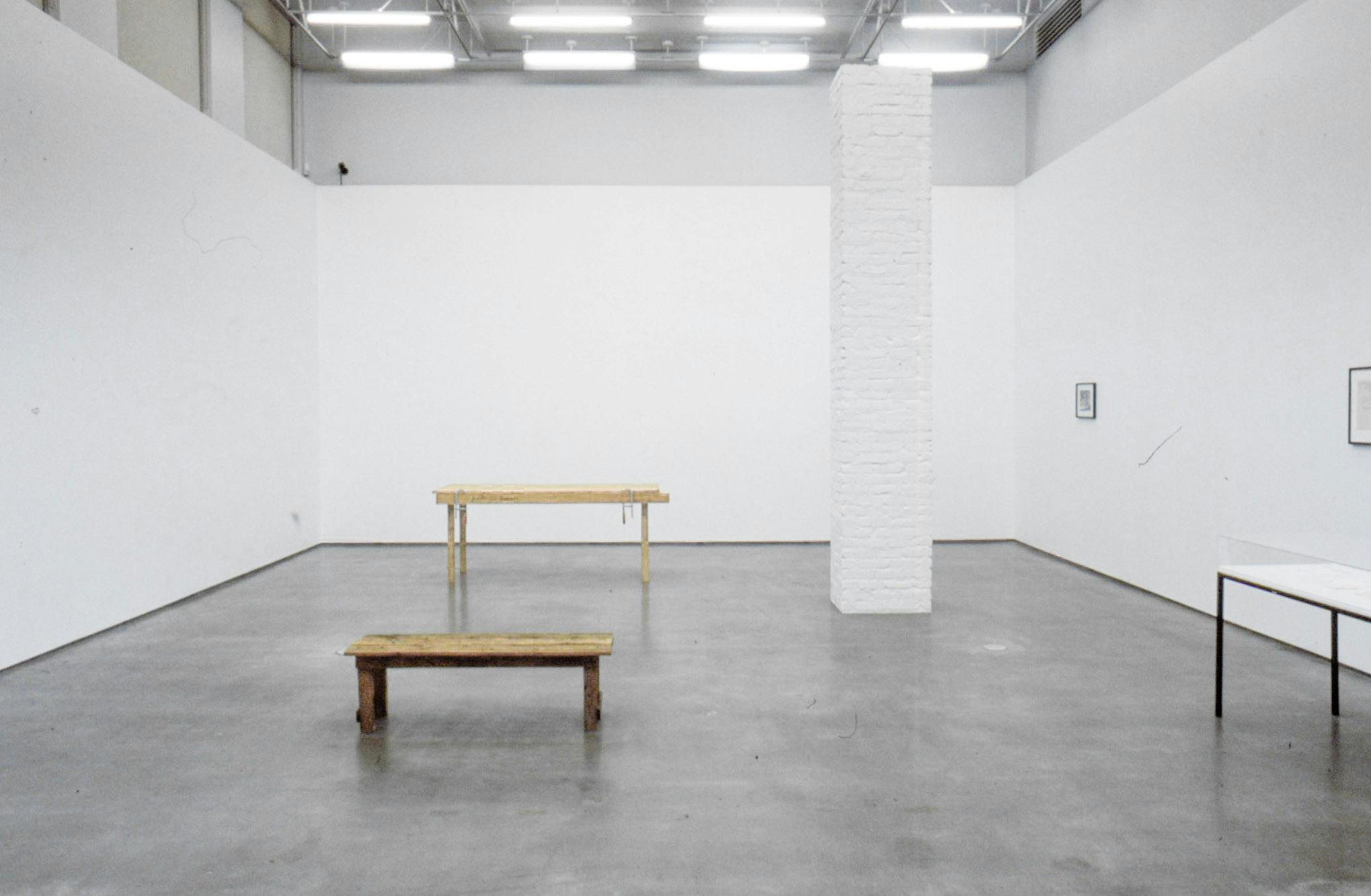 An installation image of Kirsten Pieroth’s art exhibition.  At least 7 meters tall white square pillar stands in a gallery. The pillar does not reach the gallery ceiling. A wood bench and a table are placed separately on the floor. 