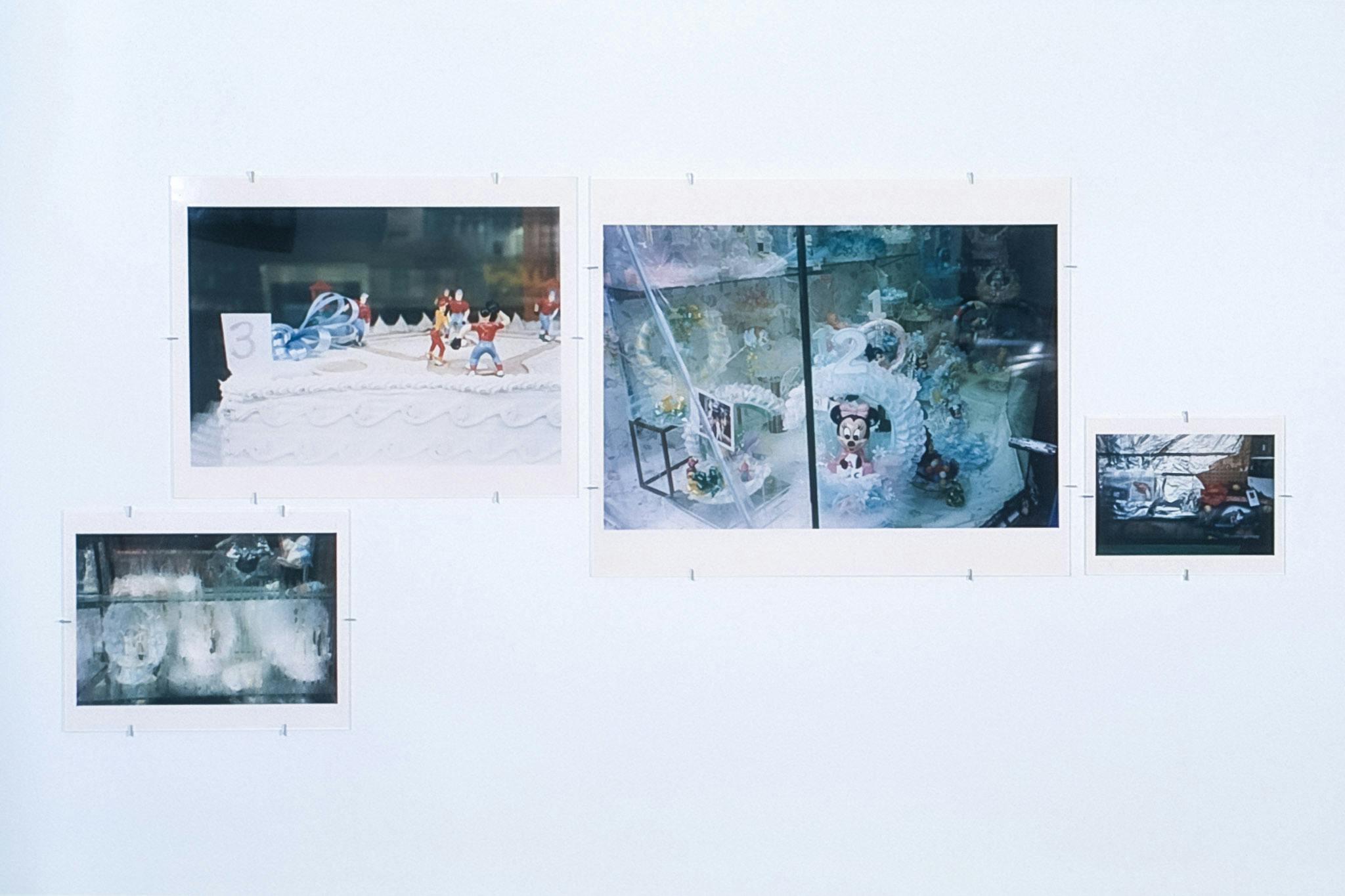 Four coloured photographs of various sizes are displayed on a white gallery wall. The largest photo among them shows small white sculptures with Disney characters and numbers on. 