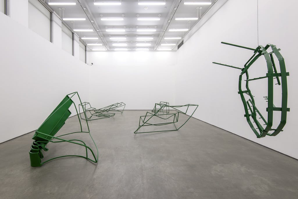 Five green, metal sculptures installed in a gallery. The sculptures resemble architectural frames and forms, distorted and bent. One of them, hanging from a ceiling, has a distorted hemisphere shape.