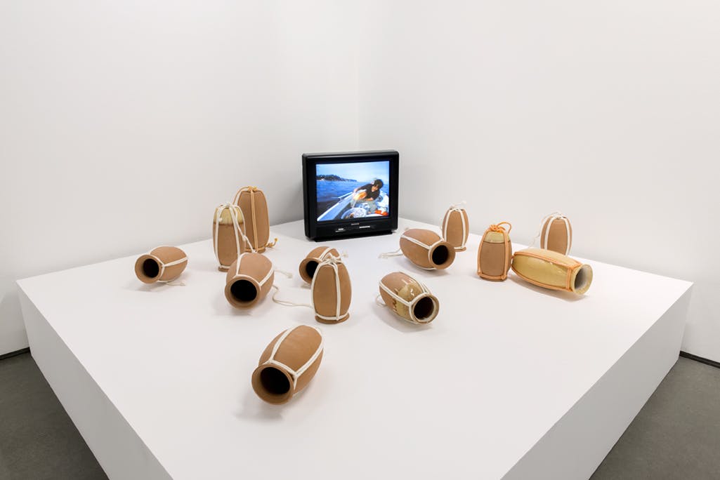 Thirteen narrow vases, which are used to trap octopuses in Japan, are placed on a large white pedestal in a gallery. A monitor plays a video of the artist using one of these traps on a boat.