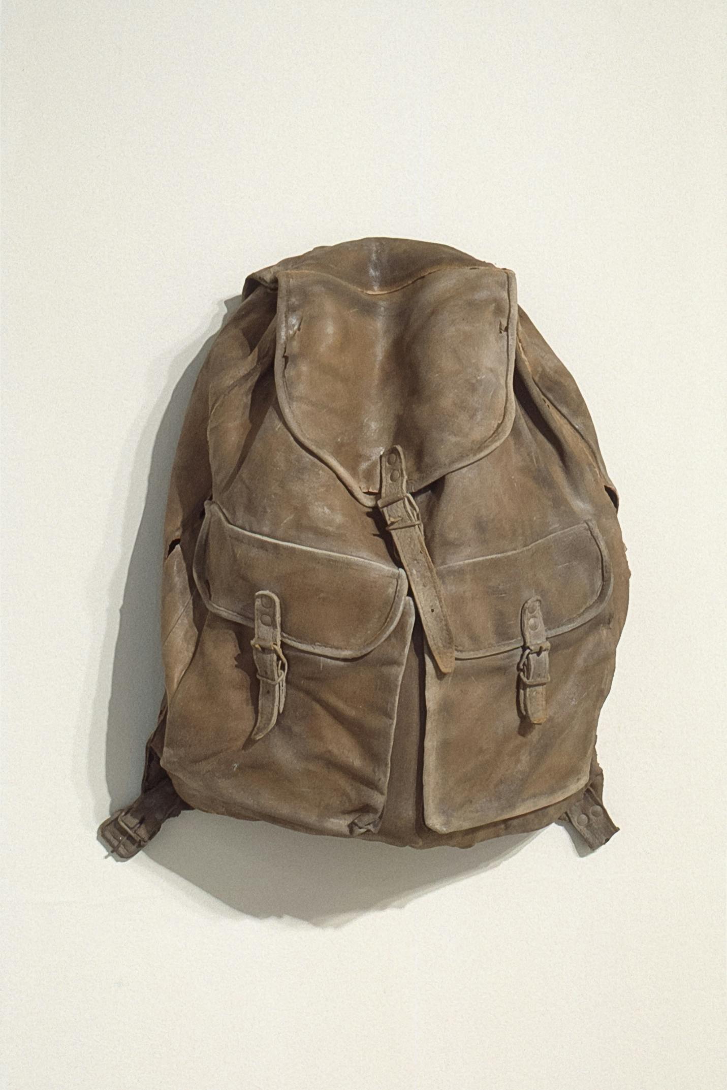 A small brown backpack with two front pockets is installed on the white gallery wall. The bag is fully closed and looks heavy. Viewers are given no clue what would be stored inside this bag.  