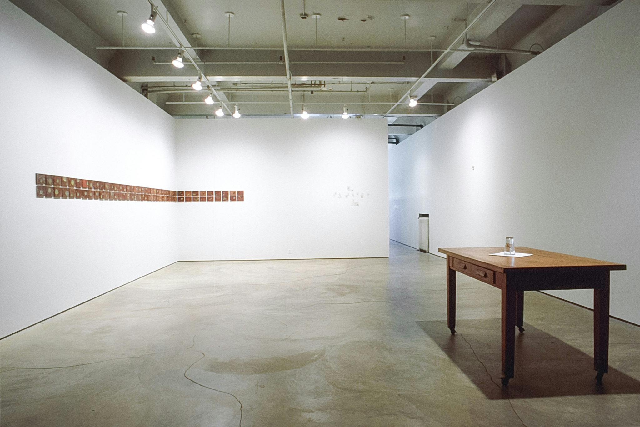 This image shows two artworks installed in the gallery. On the floor, a desk with a clear glass cup is placed. A collection of small tiles are mounted on the back wall. 