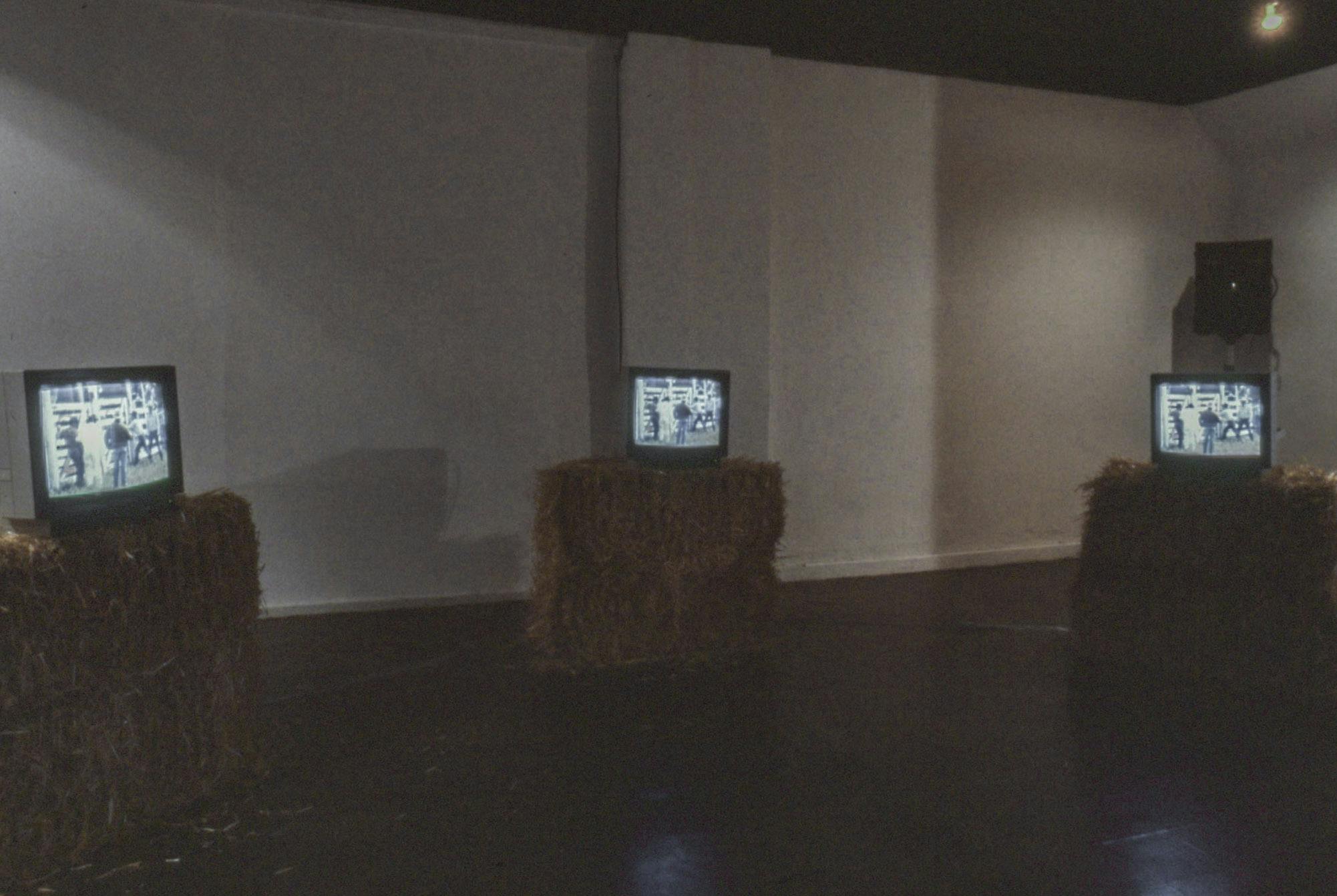 In a dark gallery space, 3 bales of hay on the floor hold a tube TV atop them. The TVs are playing the same video synchronously, showing a group of people at a barn.