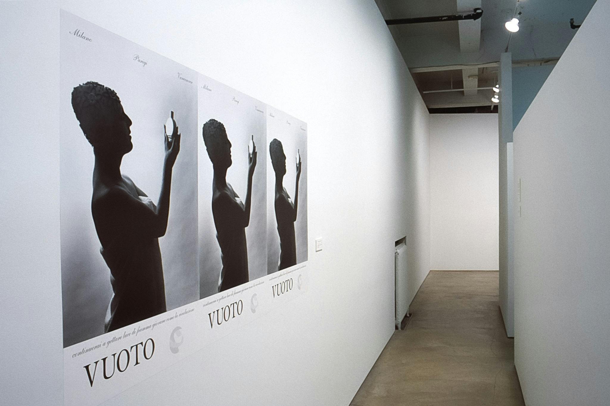Three identical posters align on the gallery wall. The poster contains a black and white photo image of a short-haired person looking at a small flat bottle. The word VUOTO is typed below the image.  