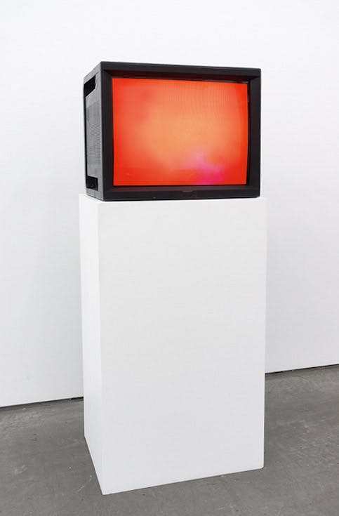An installation view of a work titled Watercolour by Ceal Floyer. A CRT TV installed on a white pedestal displays a surface of a white screen illuminated in red.