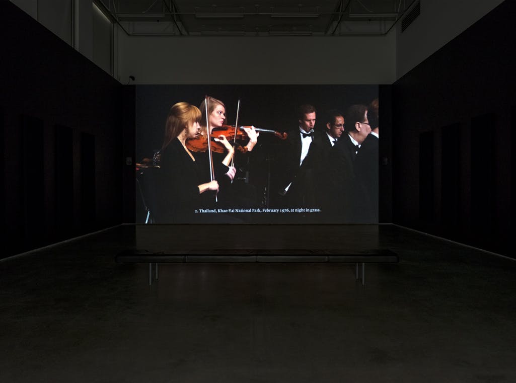 A single-channel video is projected on a screen in a darkened gallery. The video depicts musicians in black dress clothes playing instruments in an orchestra. 