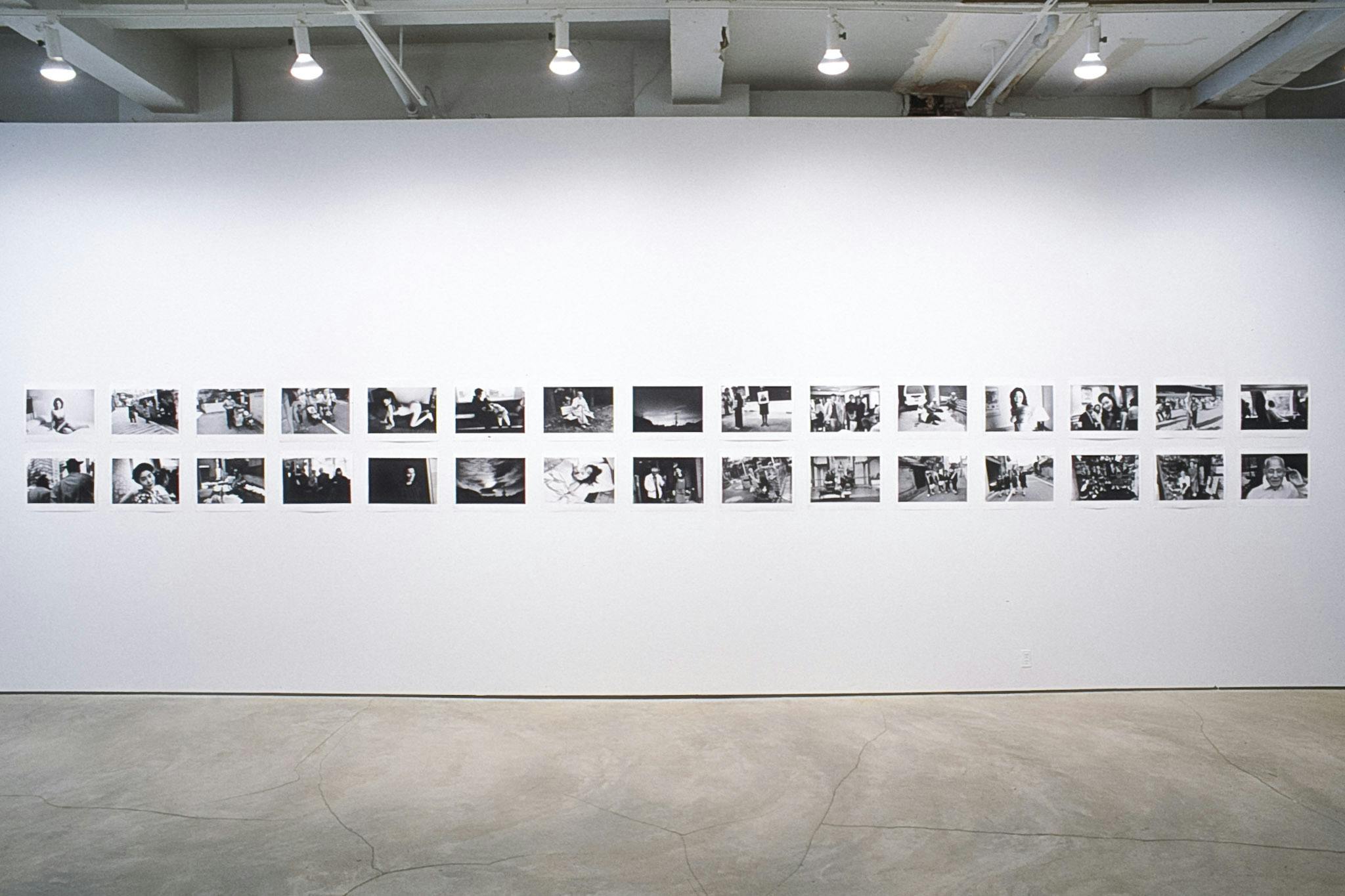 Black and white photographs are installed on a gallery wall. Many of them show human figures in various outside settings. One row contains 2 pictures and 14 rows are visible in this photograph.
