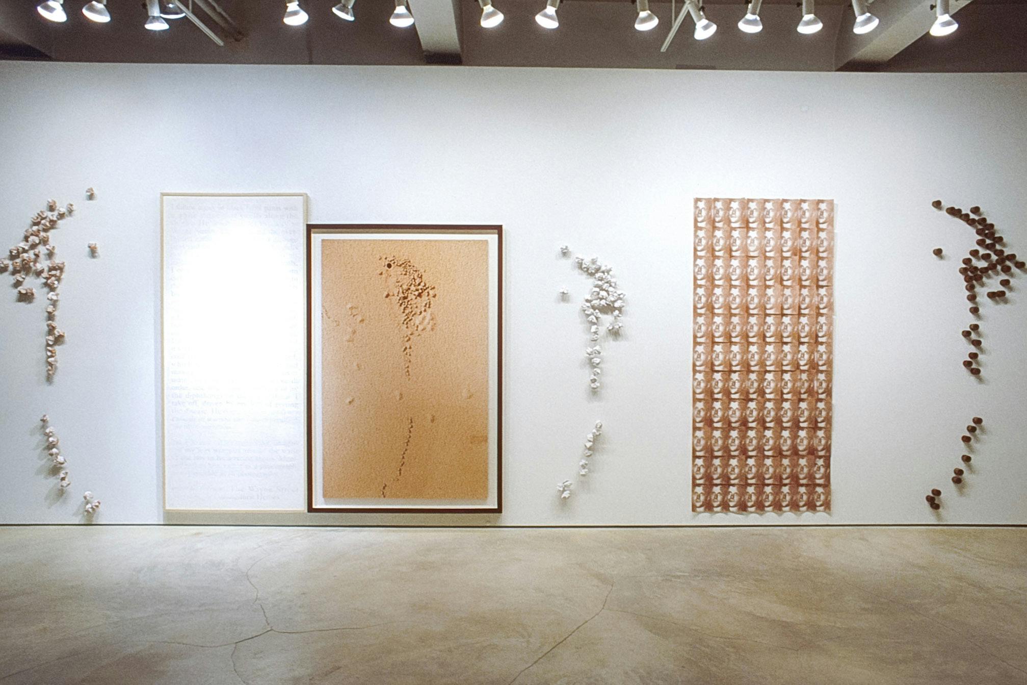 Six artworks are installed on the gallery wall. A collection of metal cylinders and two collections of folded paper are installed in between tall rectangular pictures. All artworks are in brown tones.