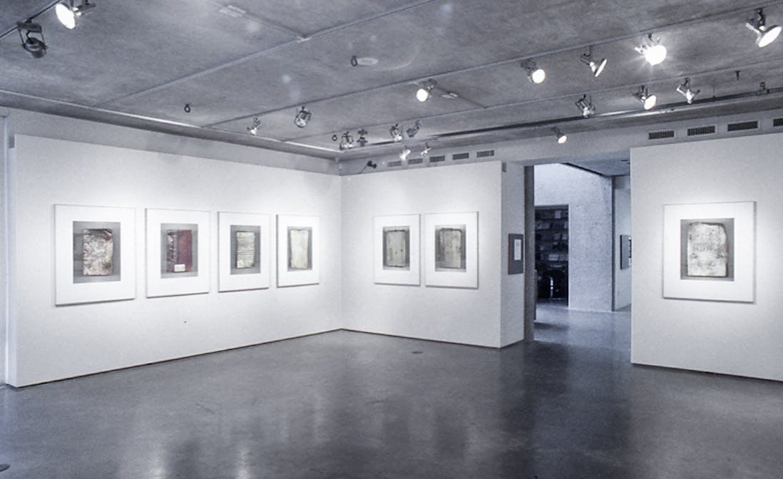 In a gallery, there are 7 framed photos and a doorway leading out into a foyer. The photos in the room are large and detailed, and show the covers of charred and crumpled books. 