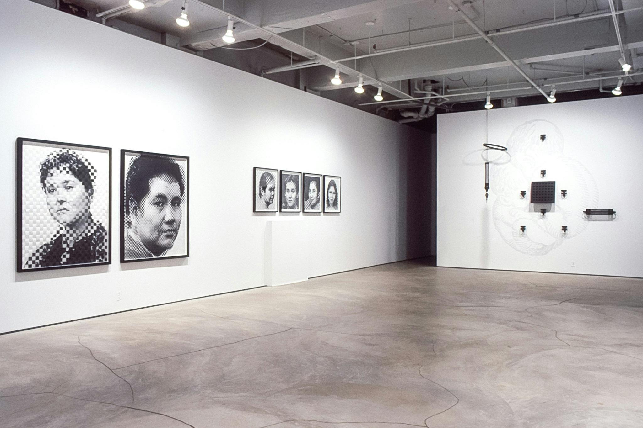 An installation view of the gallery shows artworks mounted on the walls. A group of black sculptures are on the right-side wall, and six black and white portrait photographs are on the left-side wall. 
