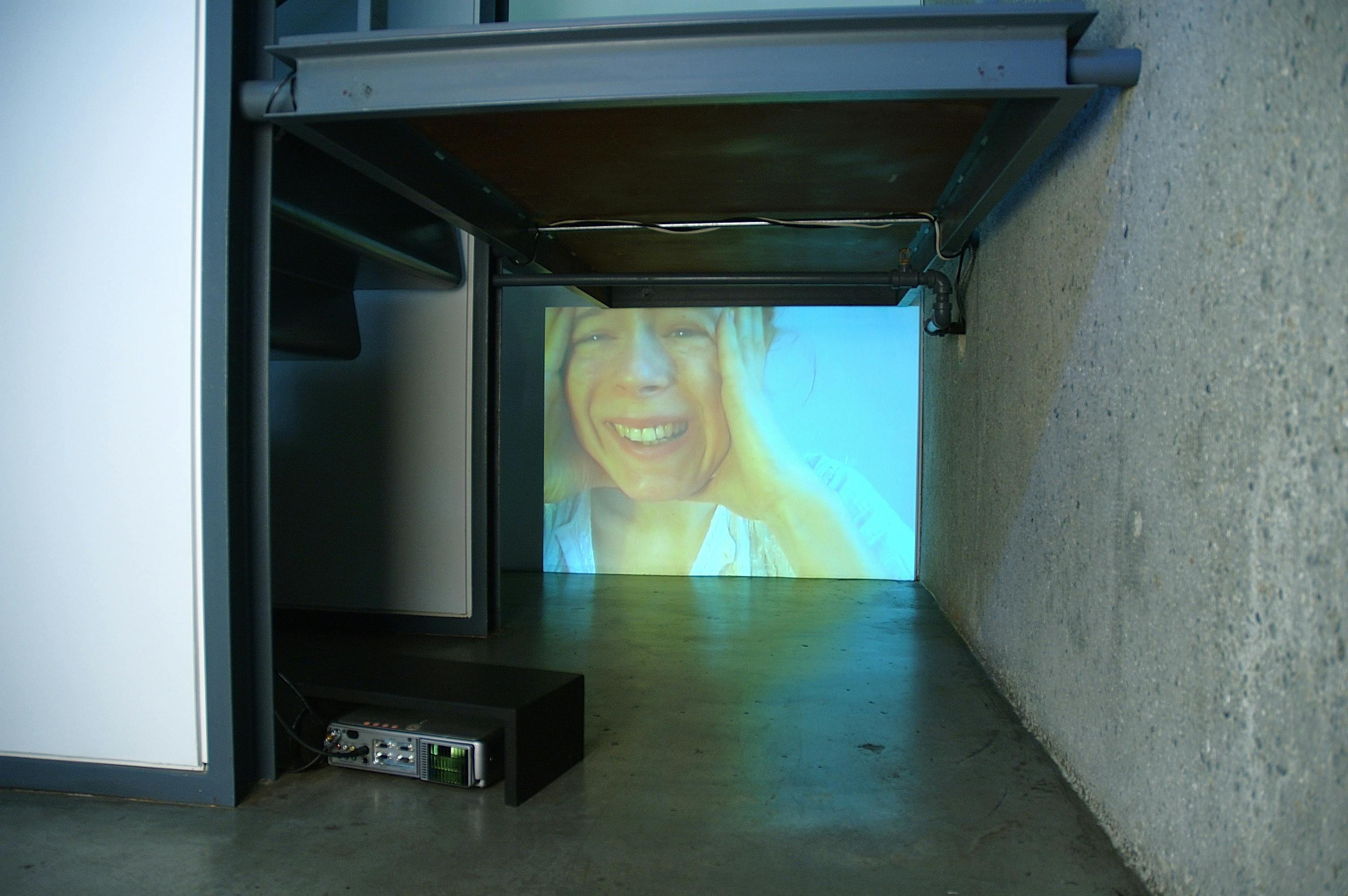 An installation image of a video projected on a wall below the staircase. The video shows an image of a person covering both sides of their head with their own hands.