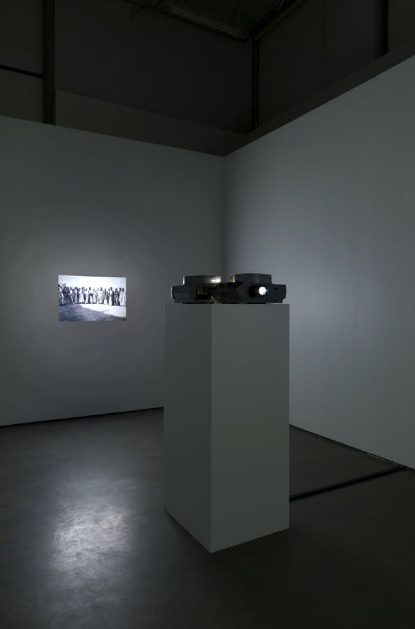 A slide projector on a plinth pointed towards a white gallery wall. The projected image is small in scale compared to the wall. It depicts a black and white image of a large group of people standing.