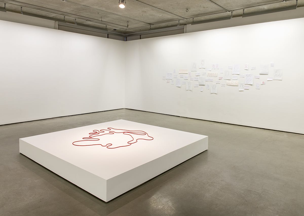 A series of drawings and a ropelike sculpture are installed in a gallery space. The sculpture is placed on a large plinth. It is a red beaded cord forming a continuous line, maplike in shape.