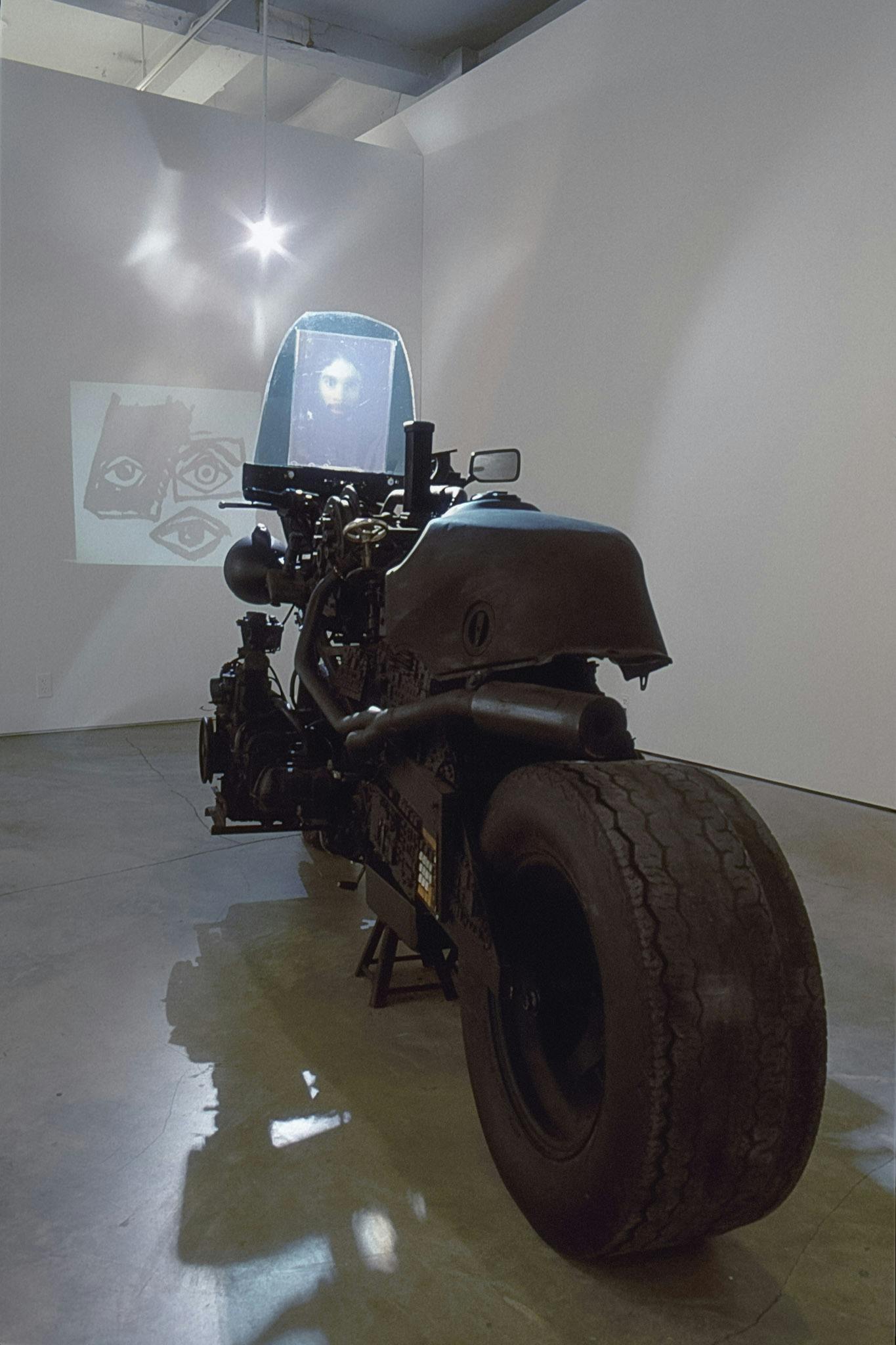 A big black motorbike is installed on a gallery floor in front of the wall, on which black and white drawings of eyes are projected. A blurred photograph of a man’s face is attached to the bike's front. 