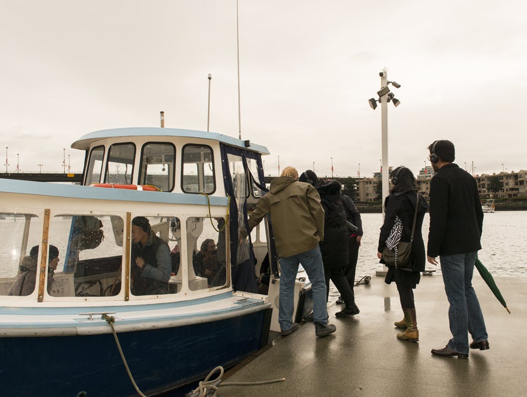 A group of people boarding a small blue boat from a concrete dock on a rainy day. A bridge and buildings are visible in the background beyond the water.