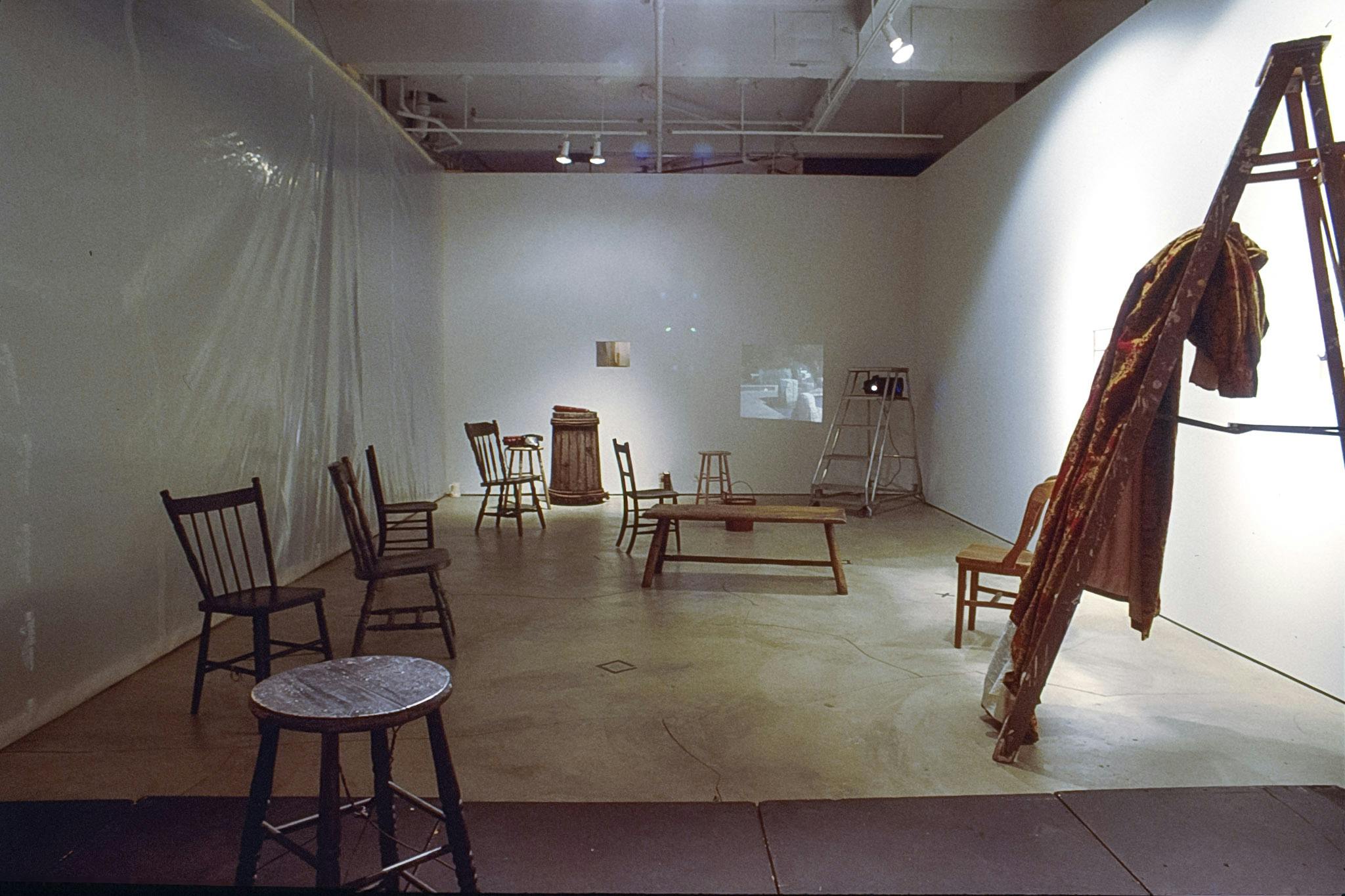 Six wooden chairs, three stools. a bench, a bucket, and two ladders are placed on the gallery floor distanced from each other. A large piece of yellow textile is hanging from one of the ladder steps. 