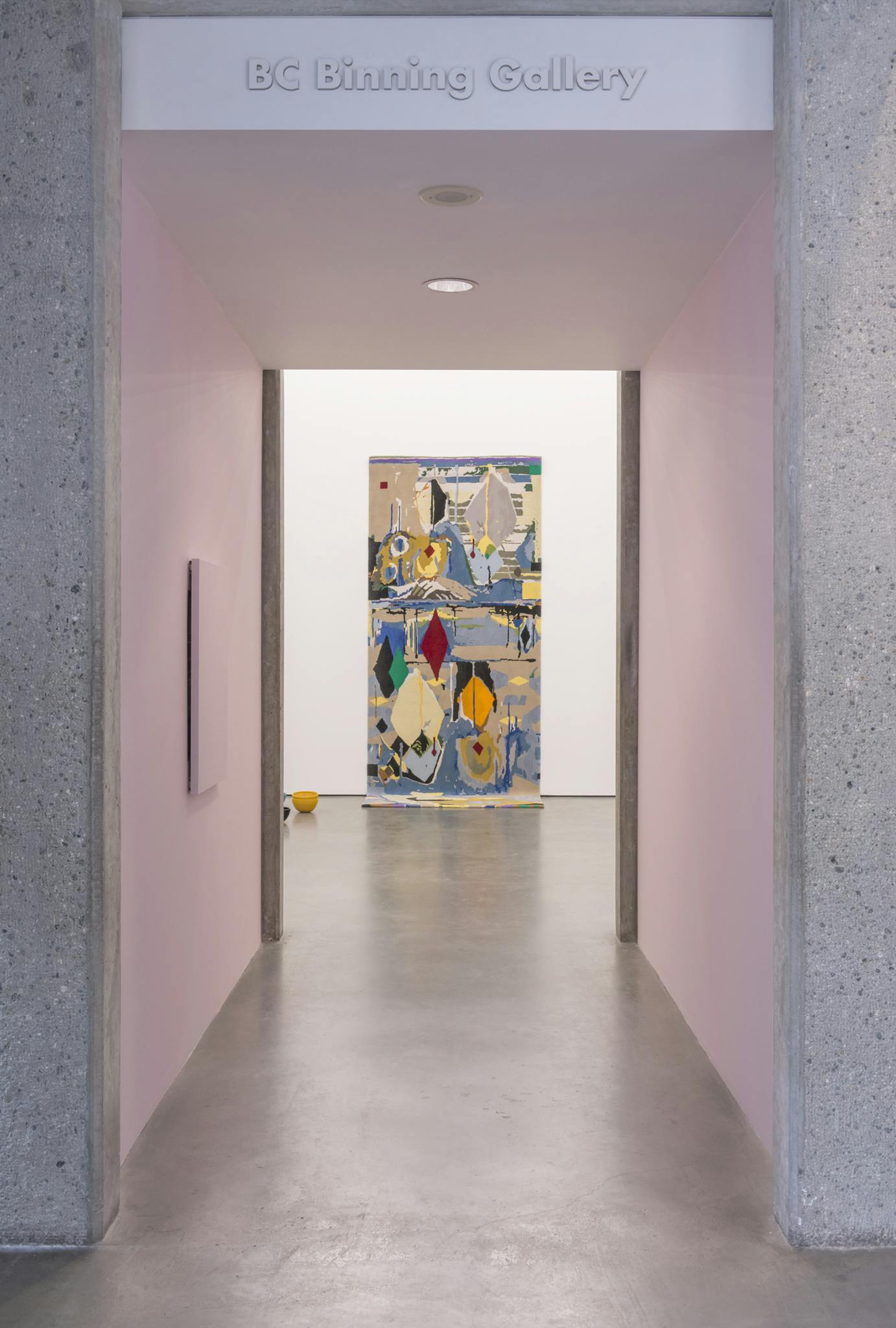 A narrow hallway to a gallery is painted light pink. A large, colourful carpet with abstract forms is mounted on the wall, visible at the end of the hallway. The carpet's end hangs to the floor.  