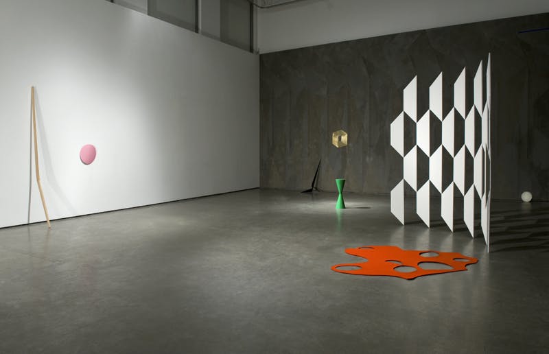 Robert Orchardson’s sculptures are installed in a gallery space. A large, fence-like sculpture of white trapezoid shapes stands in front of a dark grey wall facing a wooden sculpture across the gallery.