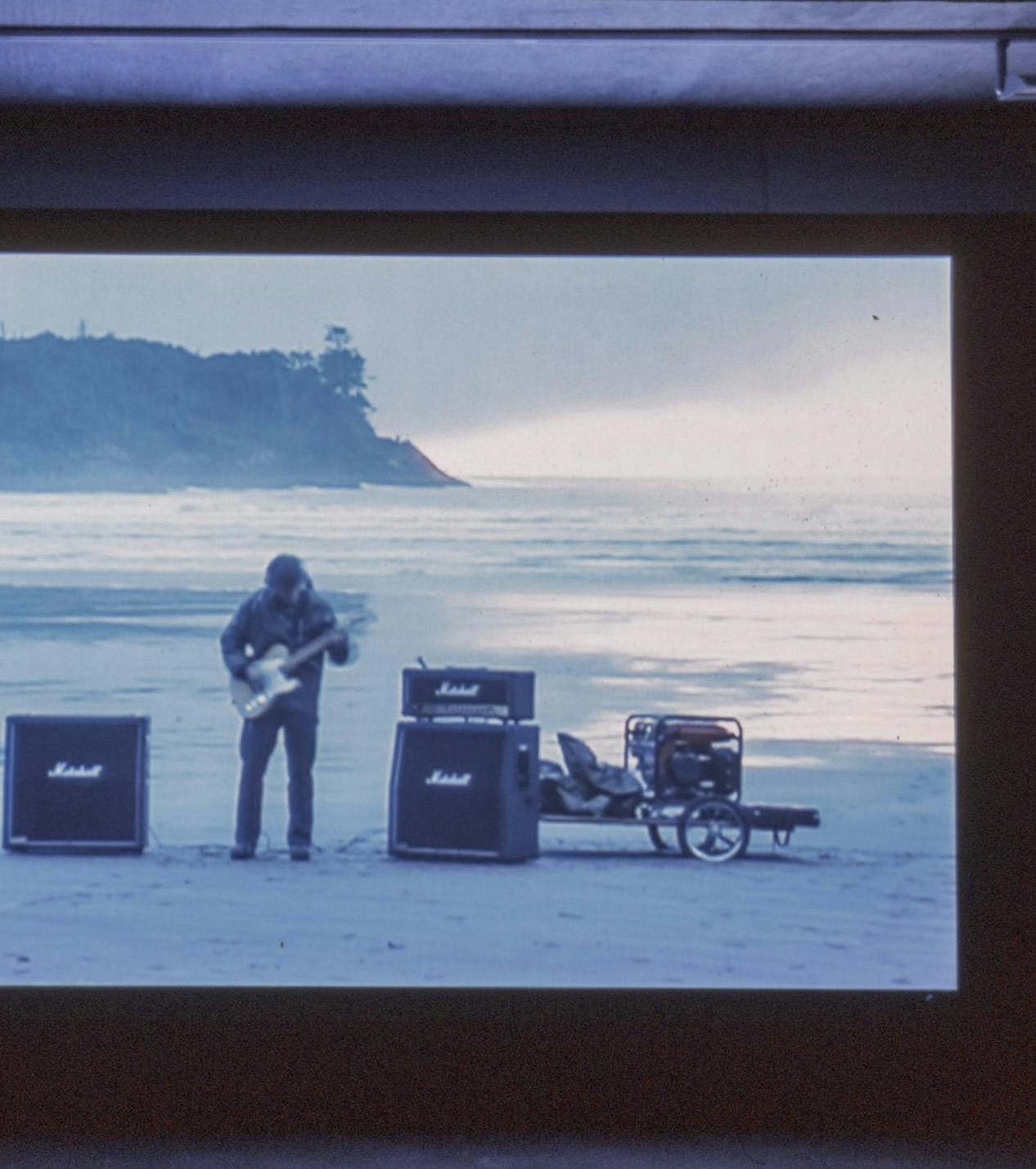 A single-channel video is projected on the wall inside a darkened gallery space. The video depicts a person playing a guitar on a seashore. The blue ocean is visible behind the person. 