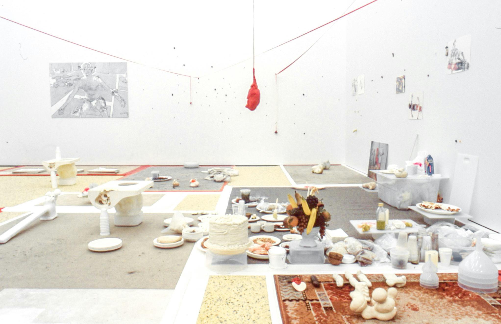 Installation image of artworks in a gallery. A variety of objects, including plastic cups, fruit, and sculptures are placed on the floor. The floor is partially covered with the patchwork of various carpets.