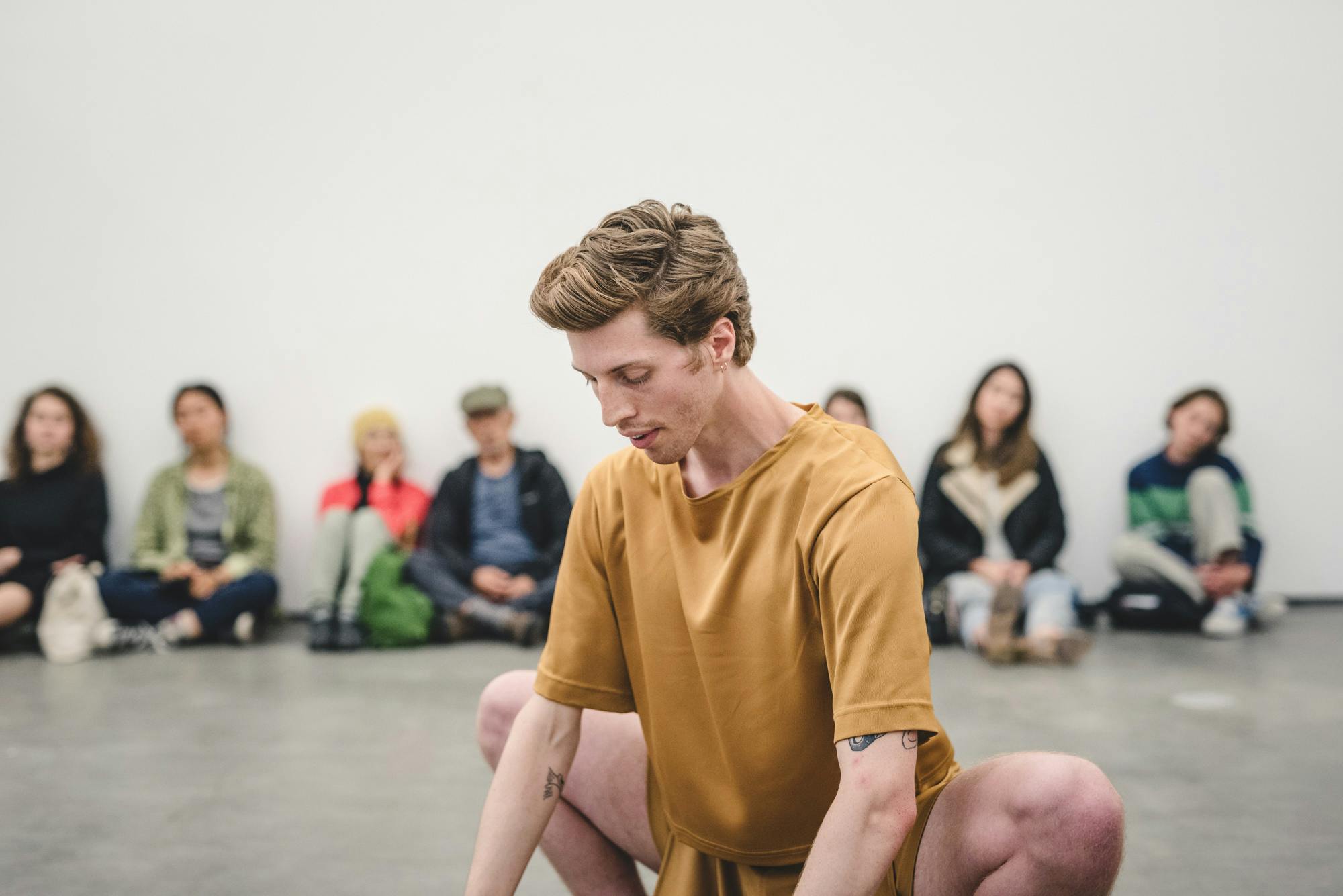 Andrew Bartee wearing a mustard coloured outfit is squatting in the middle of the gallery space. The artist’s eyes are almost closed and casted down to the floor. 