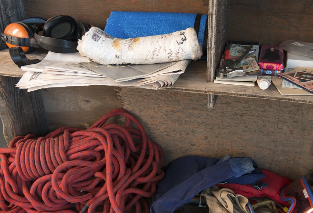 Detail image of large, rough wooden shelves installed in a gallery space. Assorted camping supplies, such as a stack of red rope, bars of chocolate, and earmuffs are placed on the wooden shelves.