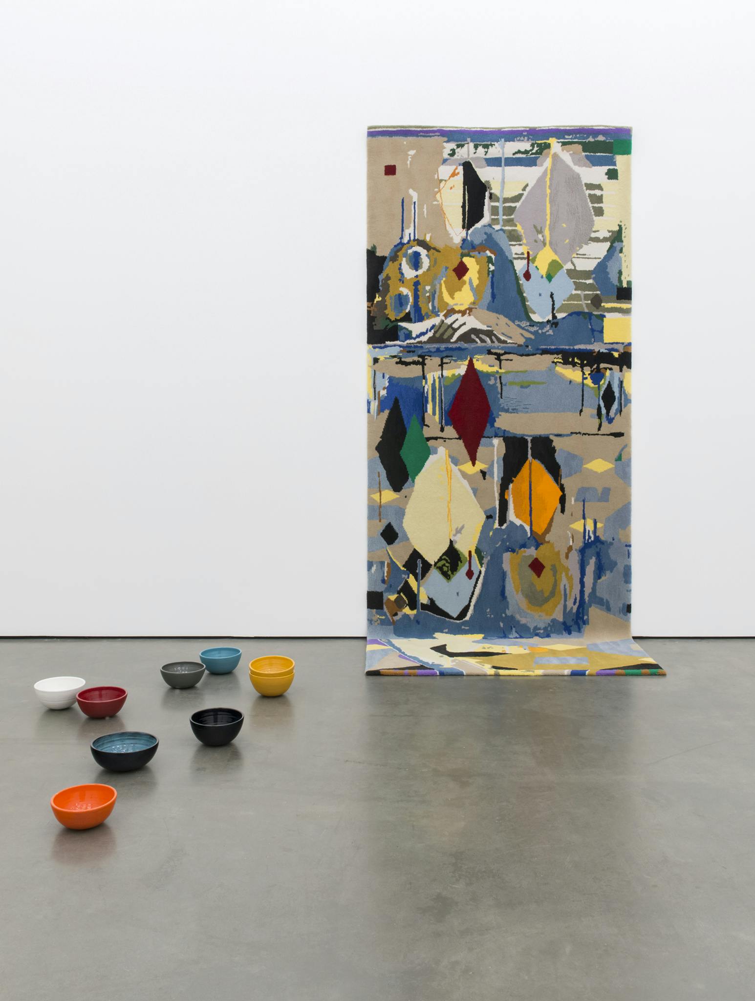 A colourful large-sized rug is installed on the white gallery wall. Eight various coloured ceramic bowls are placed close to the bottom part of the rug which is dragging on the floor.  