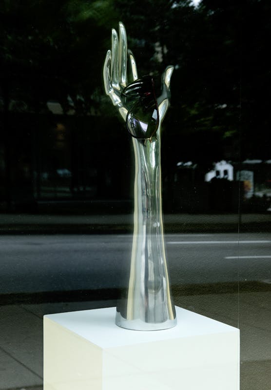 Josephine Meckseper’s sculpture installed in one of CAG’s facade windows. A mannequin arm on a pedestal loosely holds a pair of sunglasses. The arm has a silver reflective surface.