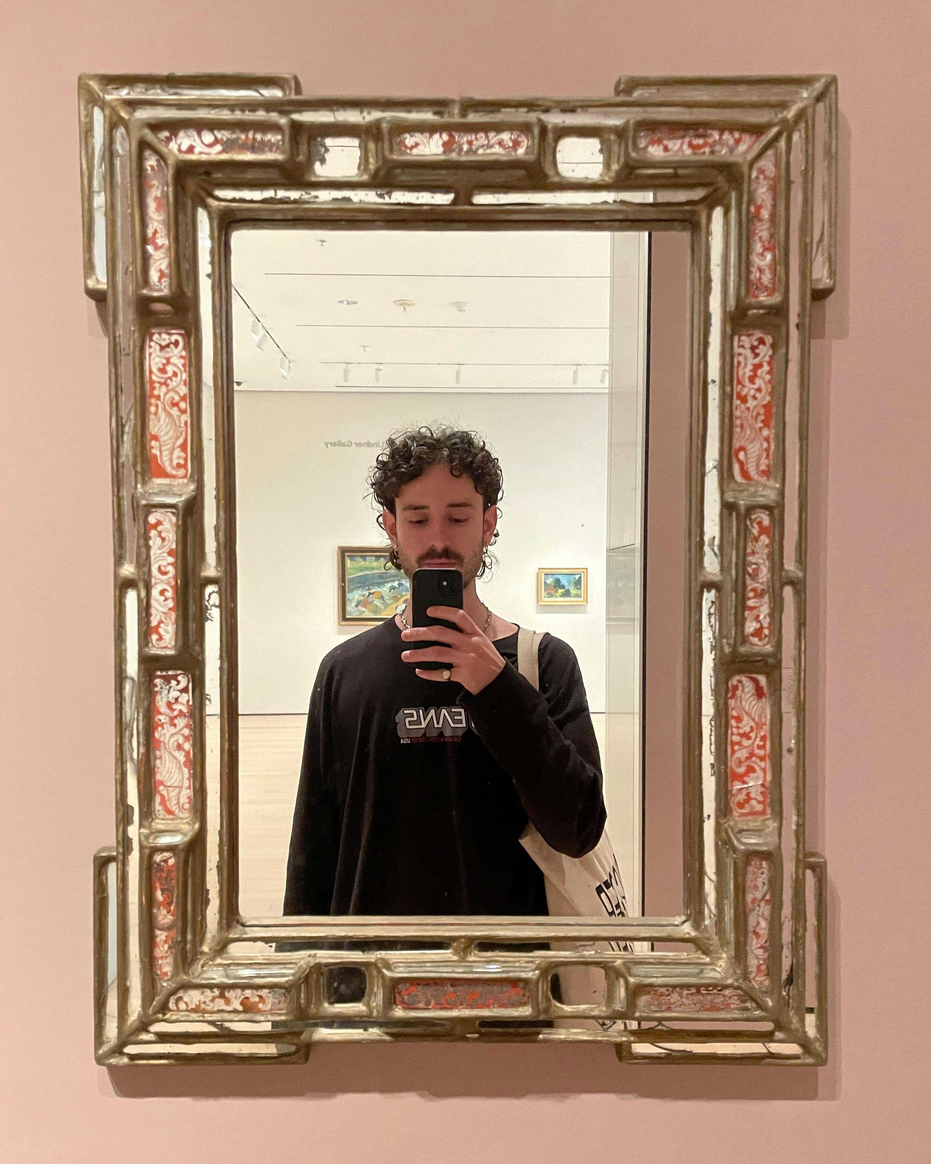 A selfie of Alex Gibson in a gallery space, shot in a mirrored artwork. They are wearing a black long-sleeved shirt.