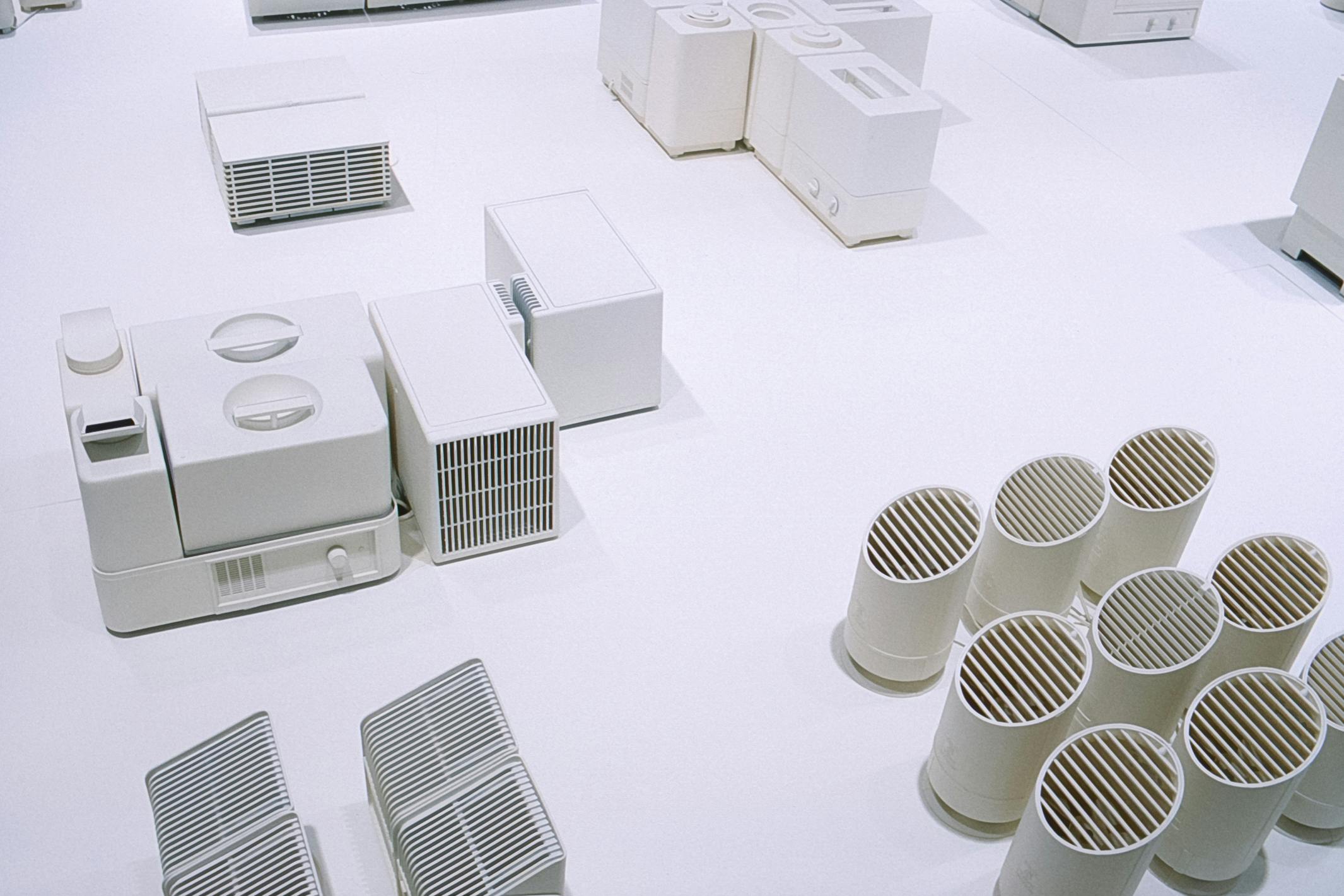 Detail of an art installation by An Te Liu. White mockups of electronic devices, many of which look like humidifiers, are placed on a white surface. 