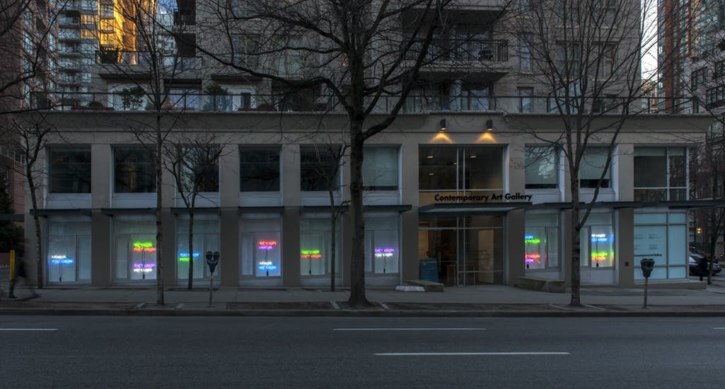 Image of Tim Etchells’ neon text works installed in CAG’s facade windows. Shot at dusk, the lights appear in various colours. 