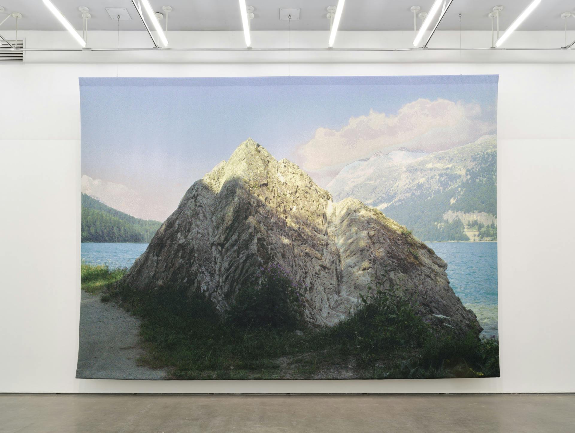 A frontal photo of a very large tapestry by Kathy Slade depicting a large rock, suspended from the ceiling in front of a white wall. 