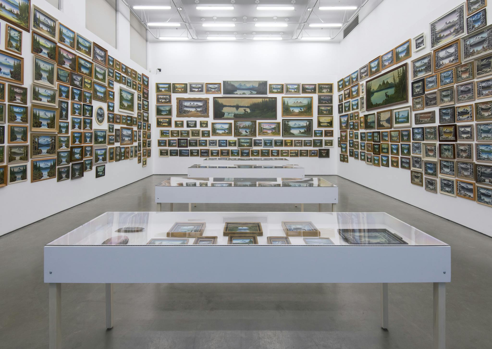 Multiple, framed paintings of similar mountain landscapes in different sizes cover the walls of a gallery in a salon-style layout. Four vitrines in the center of the gallery display more. 