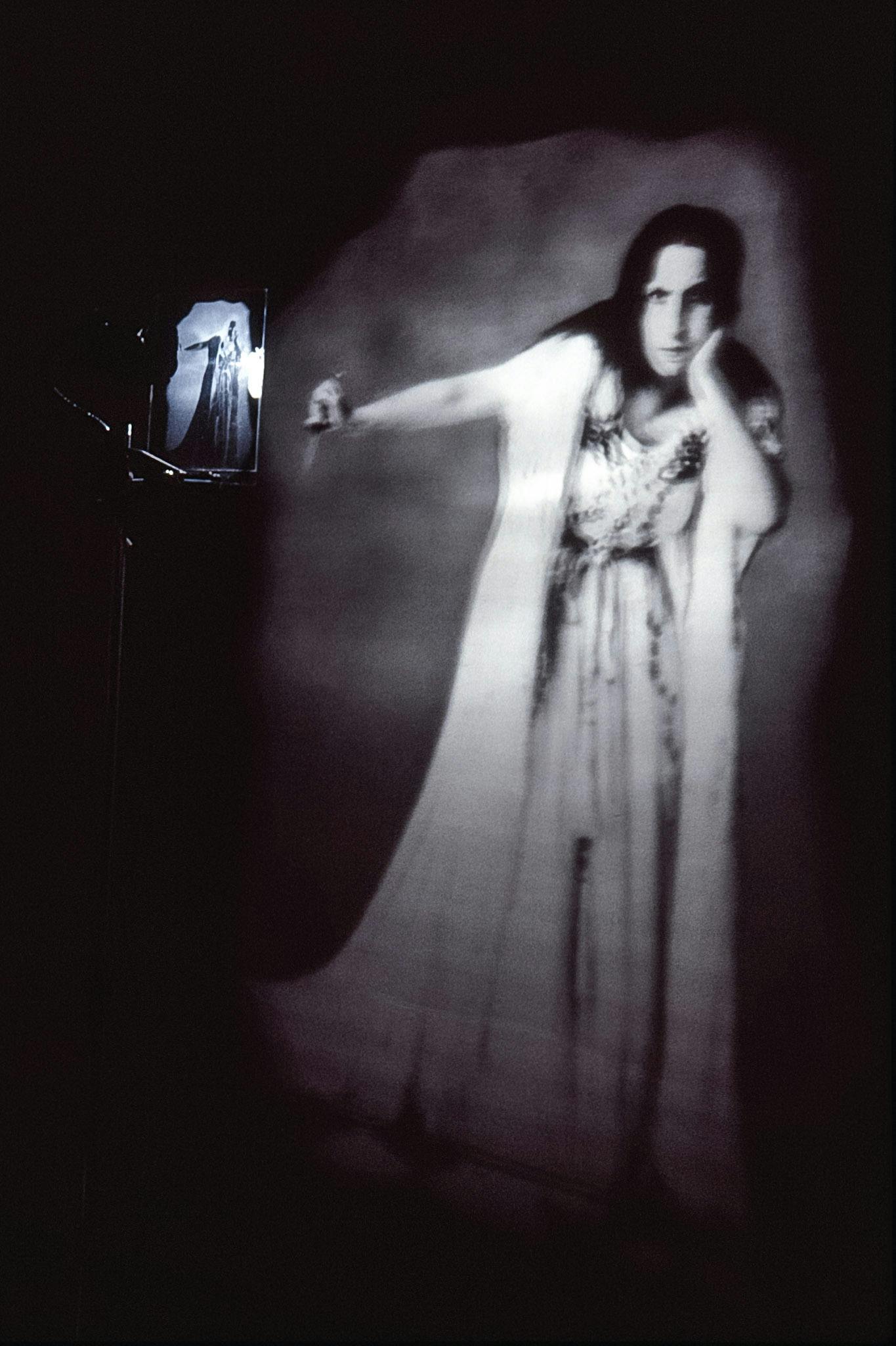 A detail of an artwork installed in a dimly-lit gallery space. A small slide installed in front of the light projects the enlarged image onto a gallery wall. The projected is a black and white image of a person in a dress. 