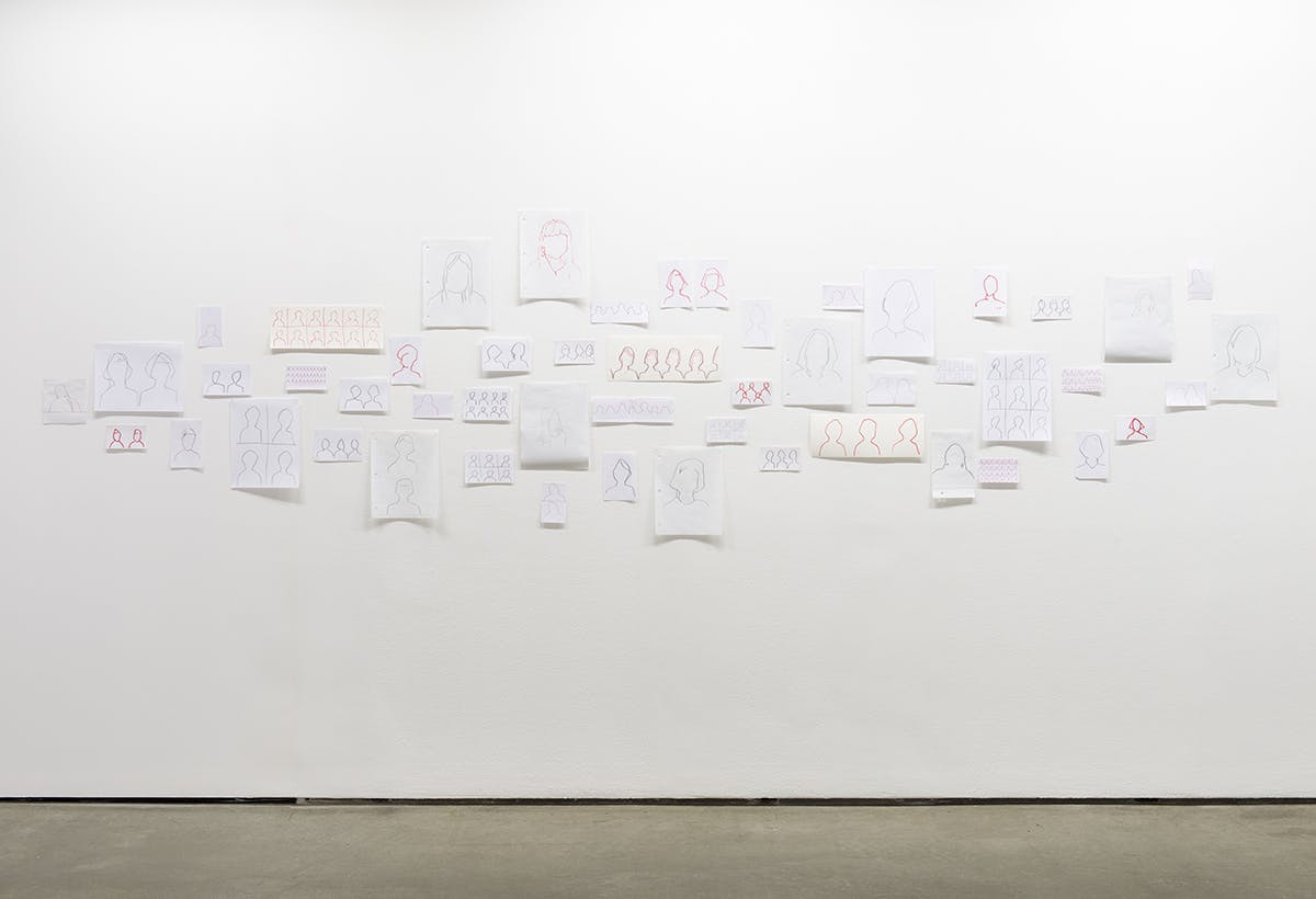 This is an installation shot of Faye HeavyShield’s drawings. Dozens of unframed drawings of various sizes and papers depict silhouettes of heads. They are hung closely to one another on a gallery wall.