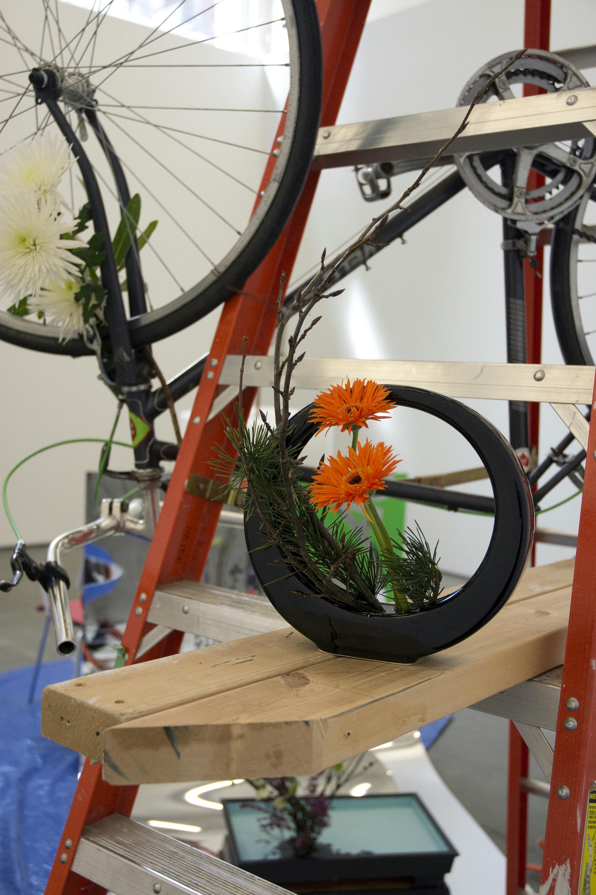 A detailed view of a sculpture installation. Orange gerberas and small twigs are arranged in a small black ikebana flower vase. The vase is situated on wood bars placed on a step ladder. 