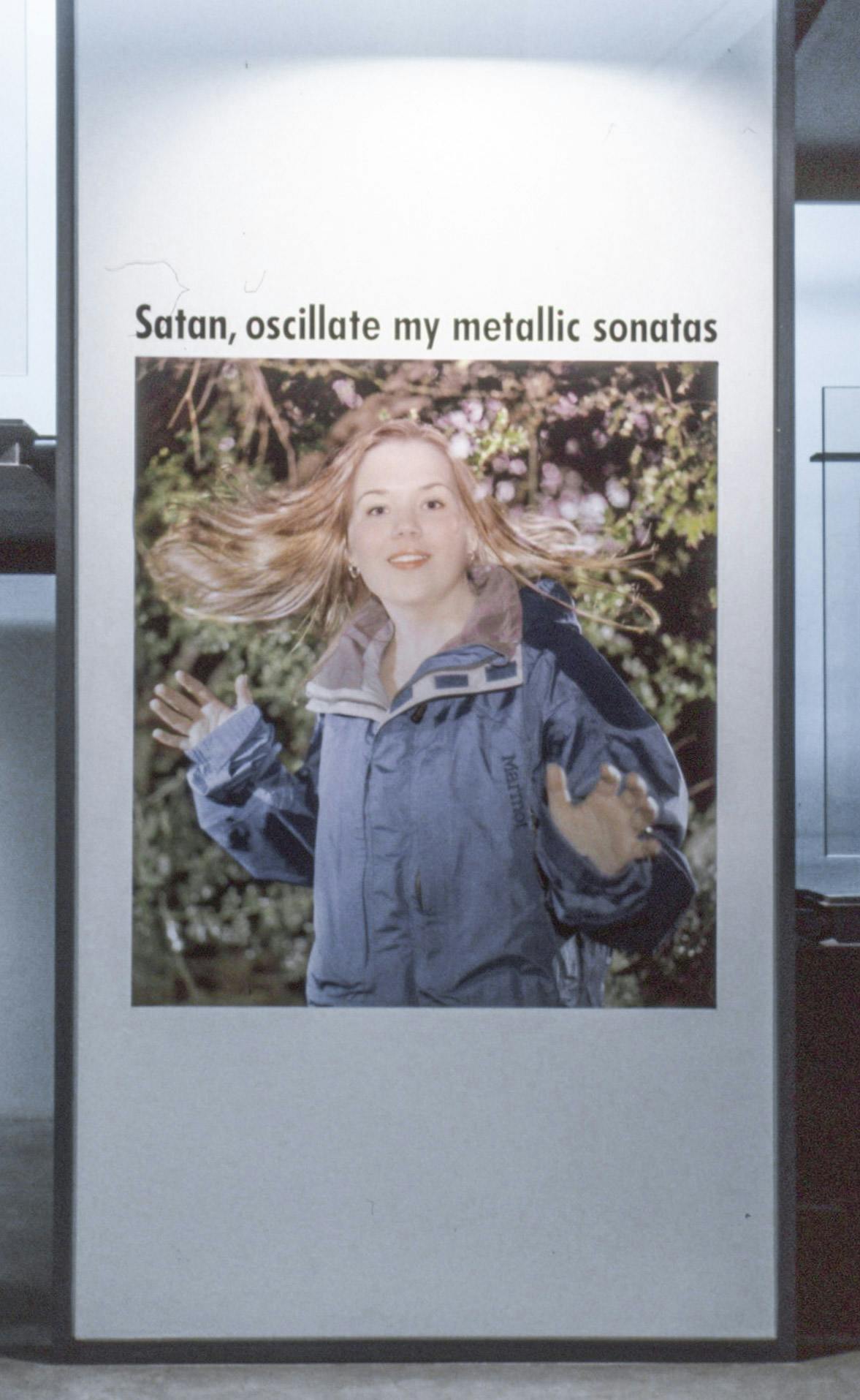 This photograph documents the entrance of the exhibition titled Satan, oscillate my metallic sonatas. A photograph of a woman standing in a forest is installed below the exhibition title. 