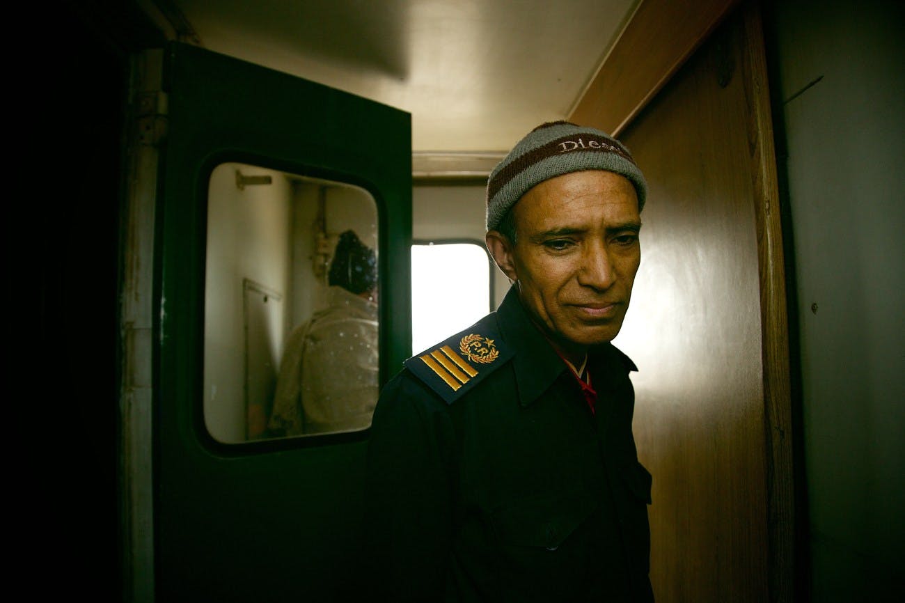A photograph of a train employee in a train vestibule. They wear a green uniform with gold insignia and a grey wool hat. They look down, away from the camera, while light from a window glows behind. 