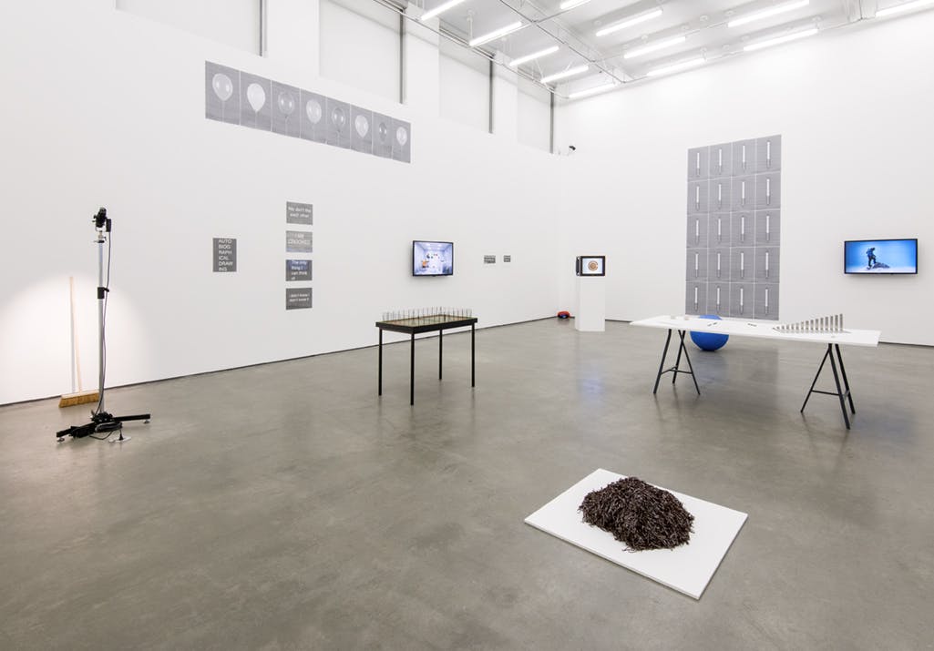 Multimedia artworks installed in a gallery: a microphone with a stand sits on the left, three monitors display video works, small sculptural pieces placed on a white table in the middle of the floor. 