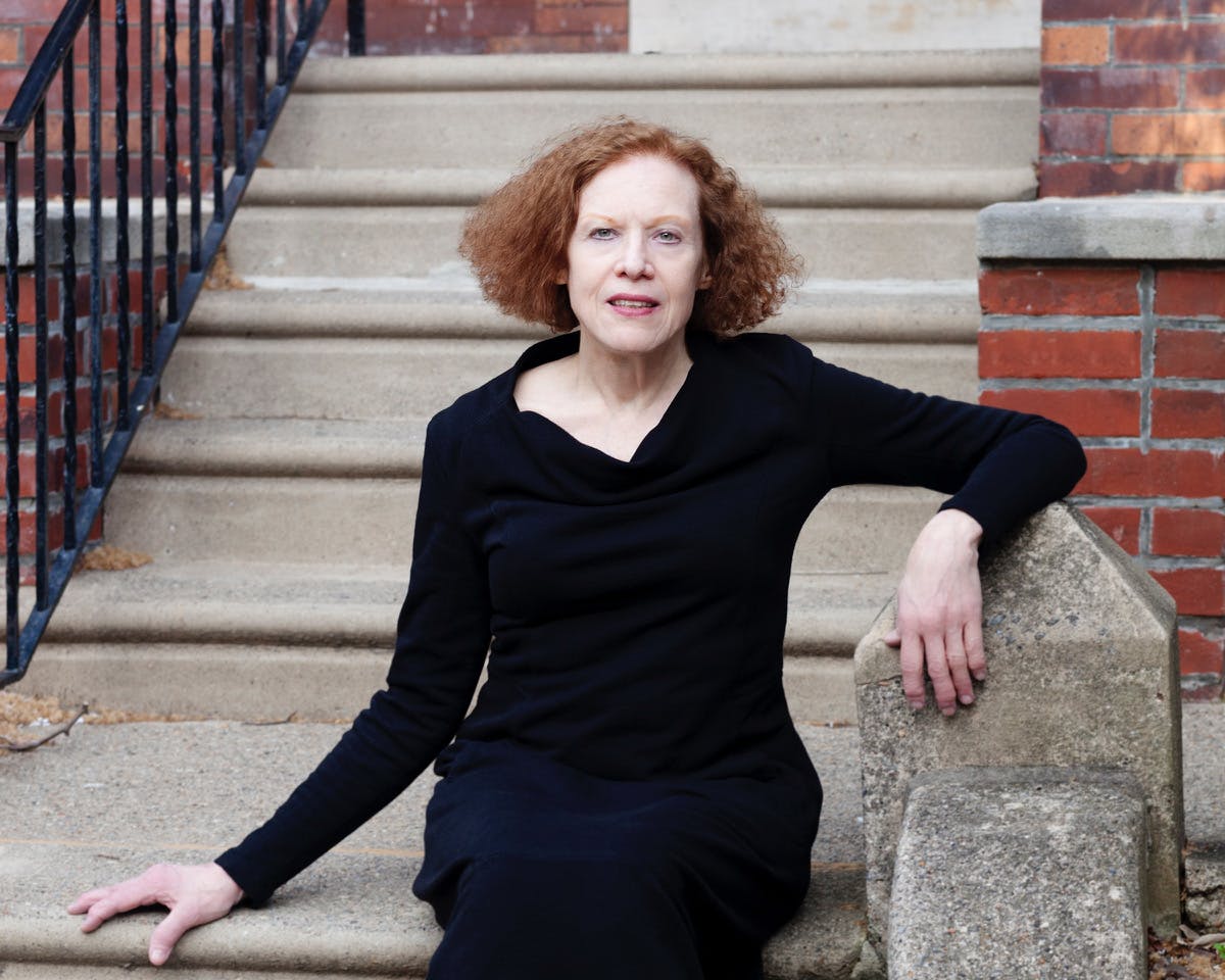 A portrait photo of Kaja Silverman wearing a black shift and sitting on stone steps outside the building entrance.