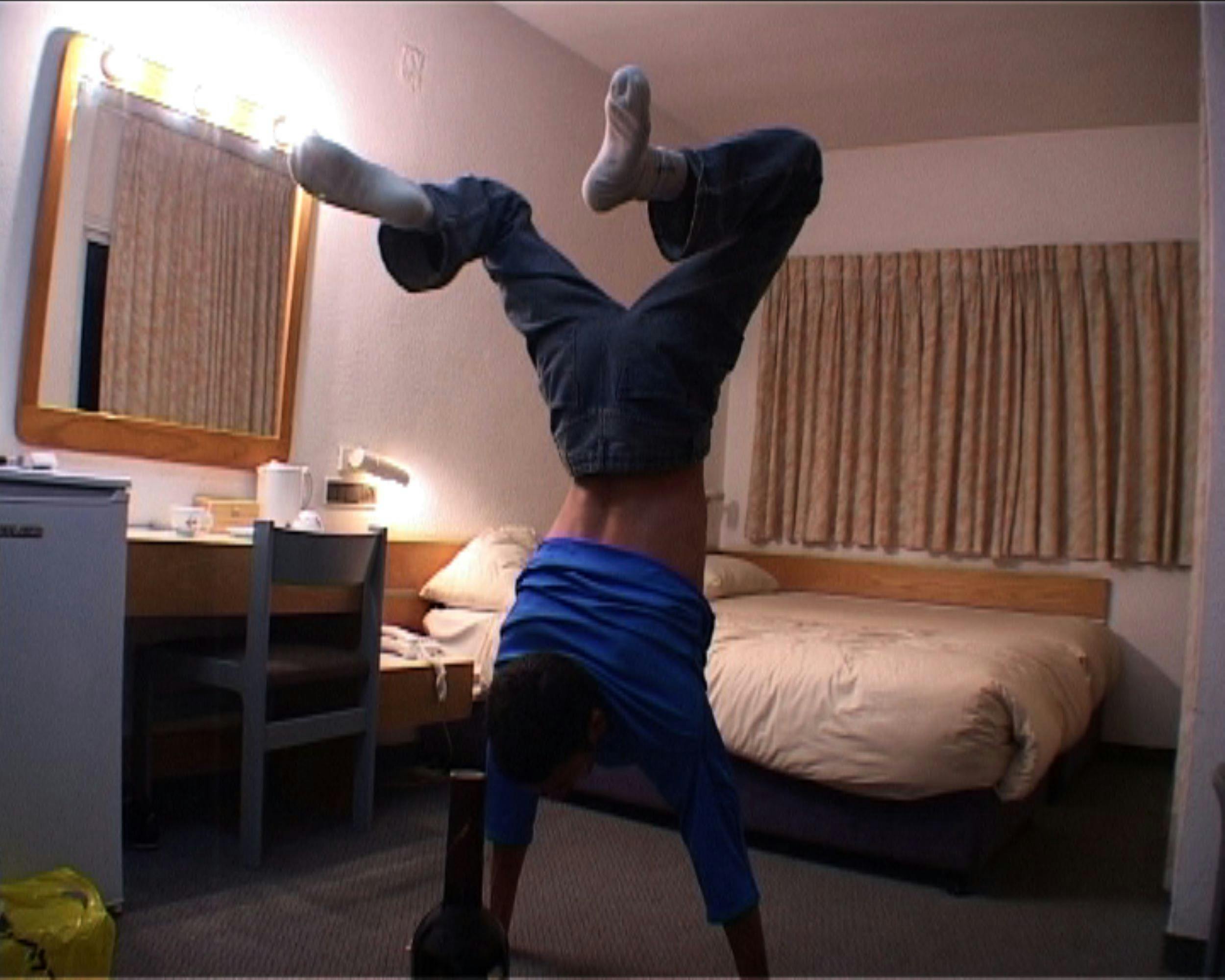 A low-fi video still of a person doing a handstand in a simple room with a beige bed and curtains, a desk, mirror, and mini fridge. A bottle is in the foreground of the image in front of the person.