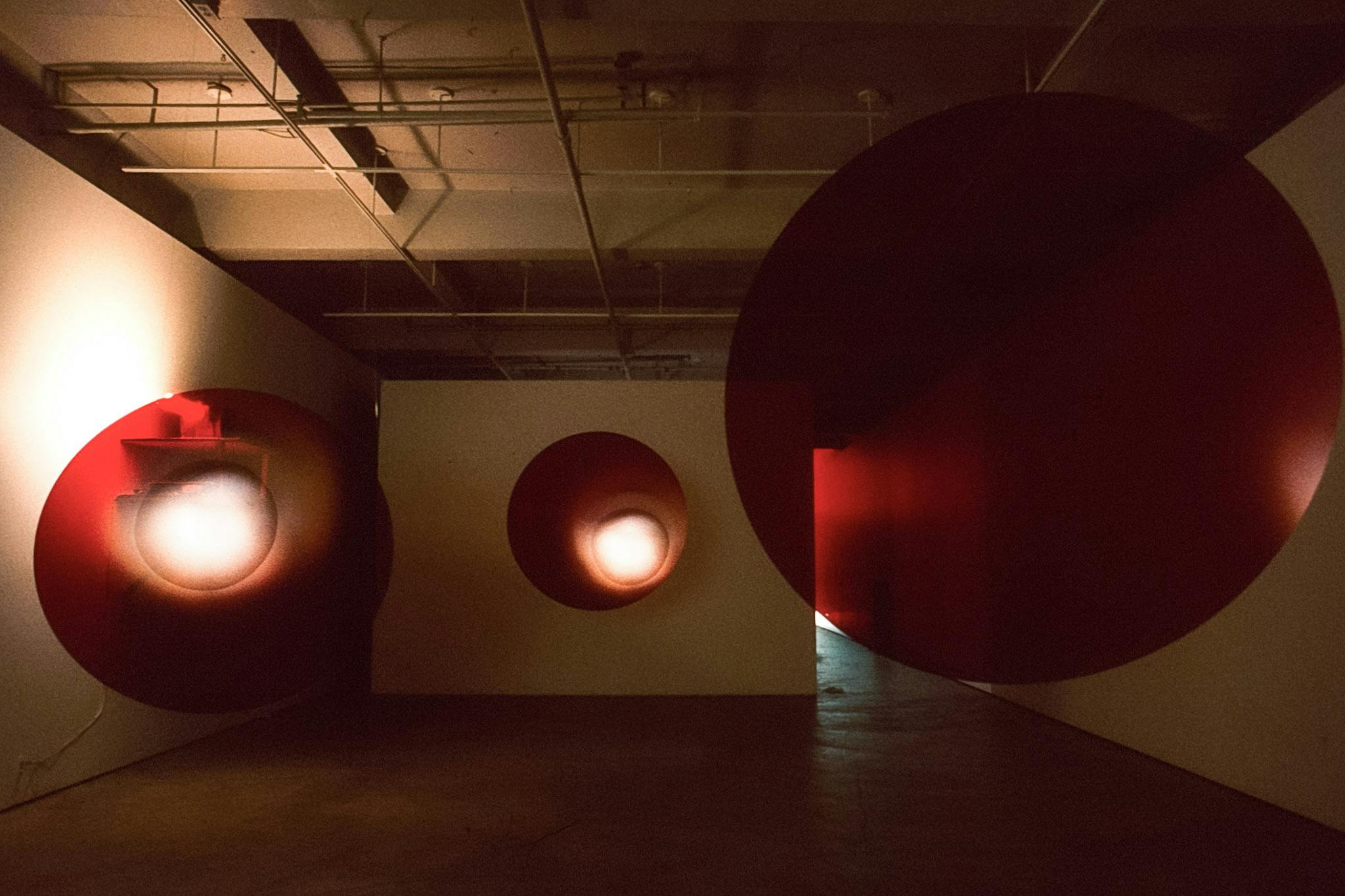 An installation view of three sculptures in the darkened gallery space. All of these sculptures, hanging from the ceiling, are large-scaped, thin, and have a red circular shape.  