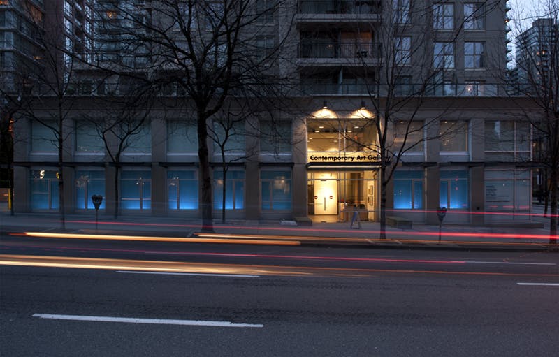 Scott Massey’s works are installed across CAG’s facade windows at dusk. Blue fills the window vitrines with white light glowing at the bottom. Cars and pedestrians pass by in front of the windows. 