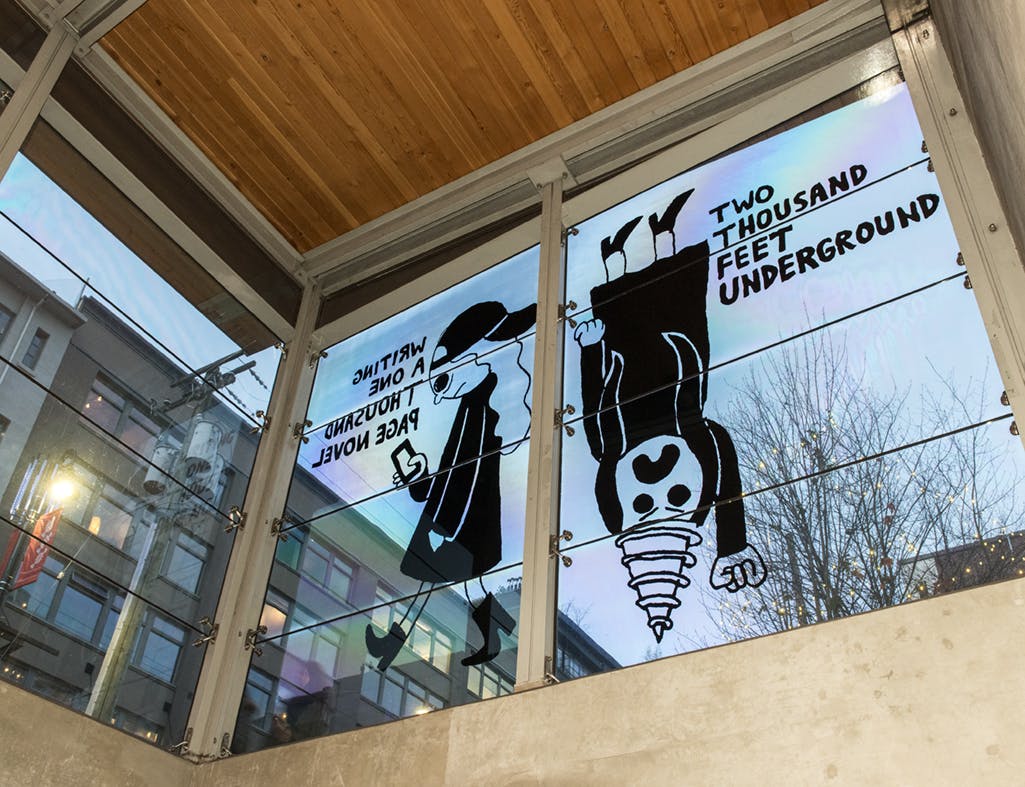 A graphic illustration of two figures printed in black vinyl on the glass face of a train station. One is walking and the other is upside down with a drill-like head. Bold text is on either side.