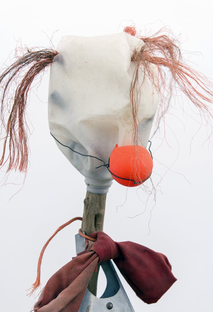 A sculpture resembling a clown. A plastic jug from which orange fibres emerge is fastened upside down on a wooden stick with a red bow tied to it. A bright orange clown-like nose is held on by wire.